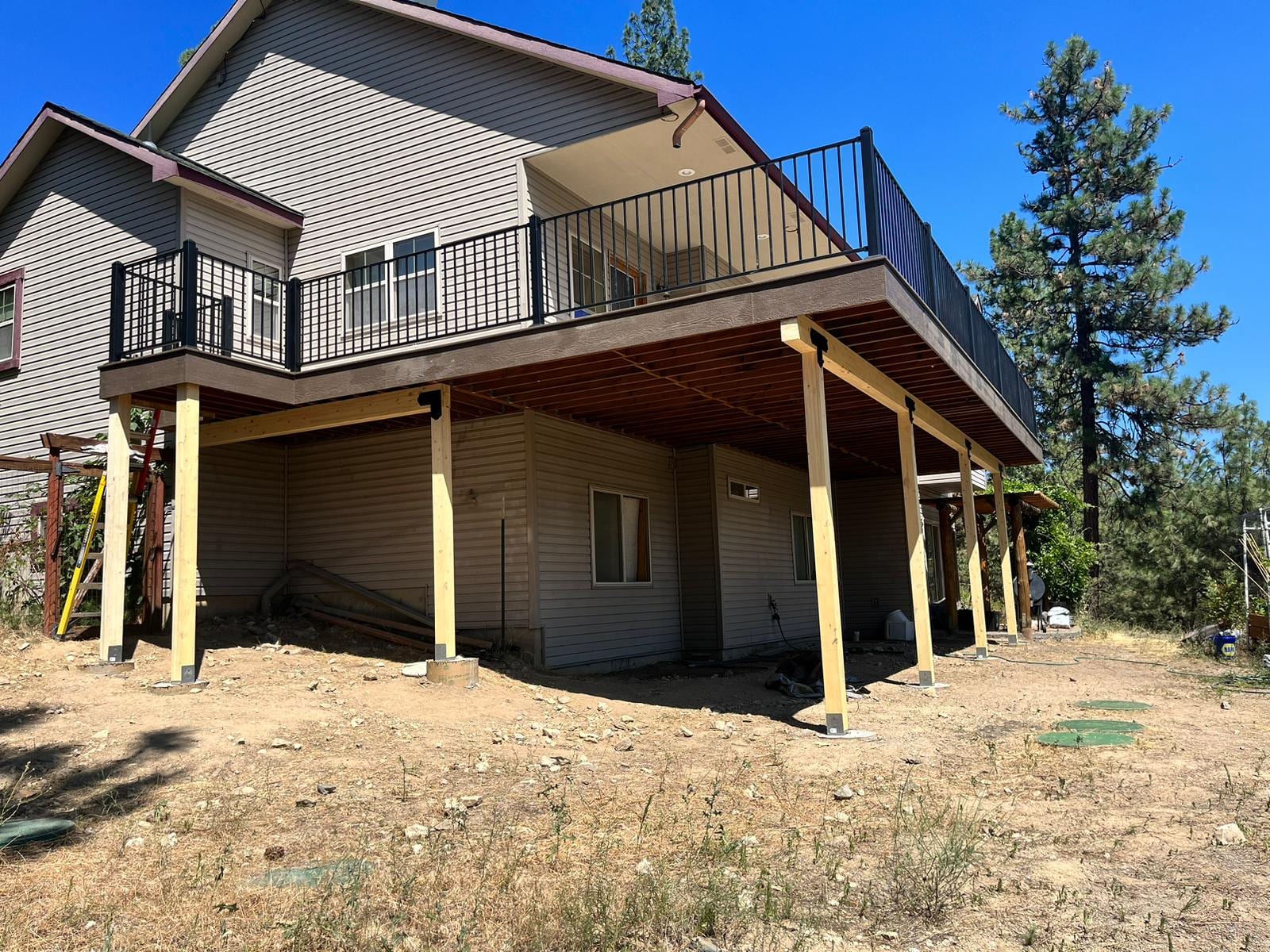 Elevated deck with black railing on yellow-stained posts, bare dirt yard and tan siding house