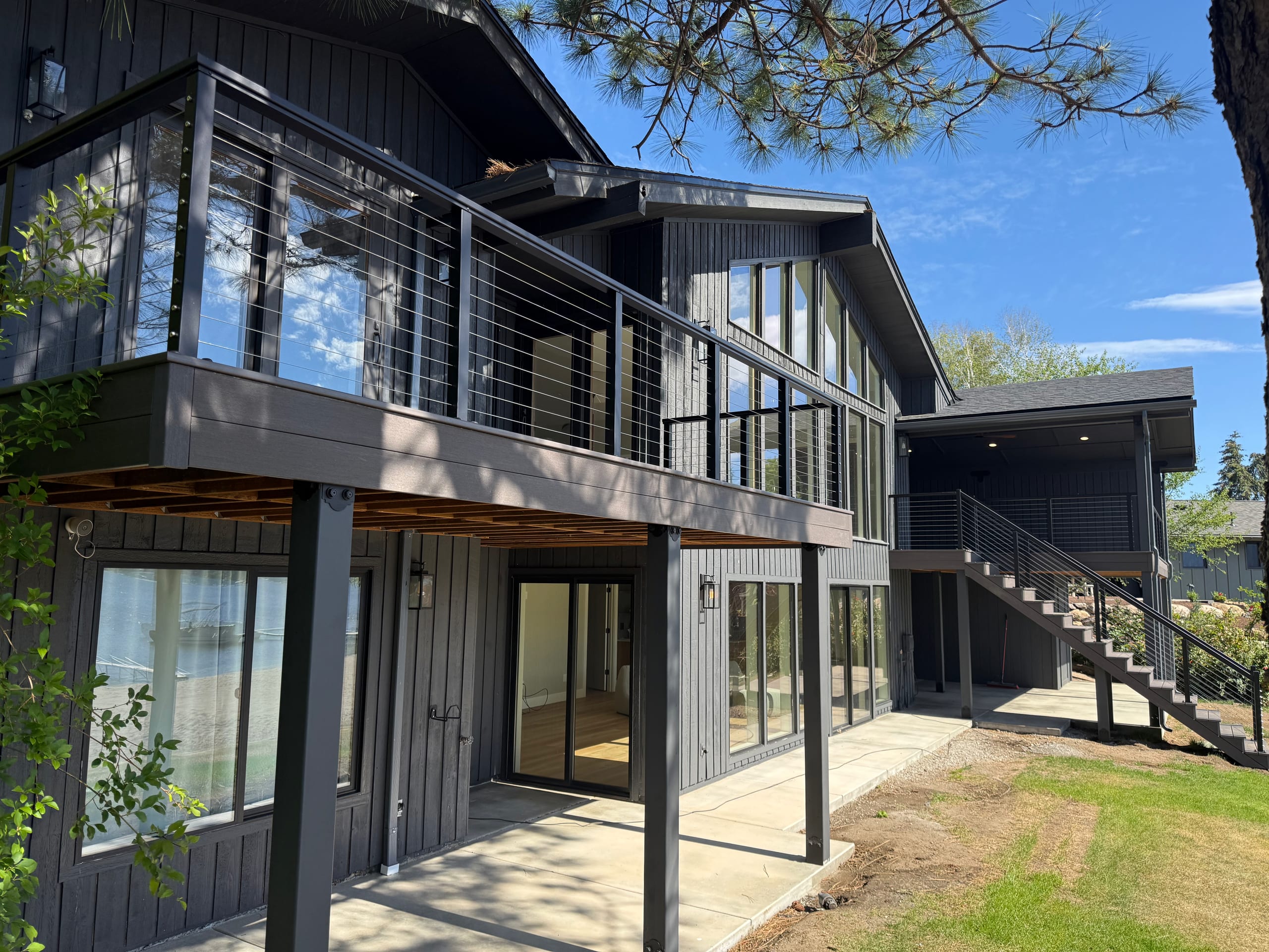 Elevated deck with cable railing wrapping dark modern house with tall windows, stair at right, sunny