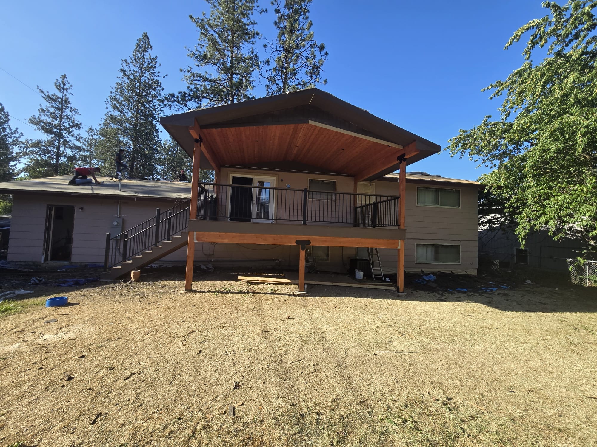 Elevated deck with cedar-ceiling hip cover and black railing on a split-level house, bare dirt yard, pines and blue sky