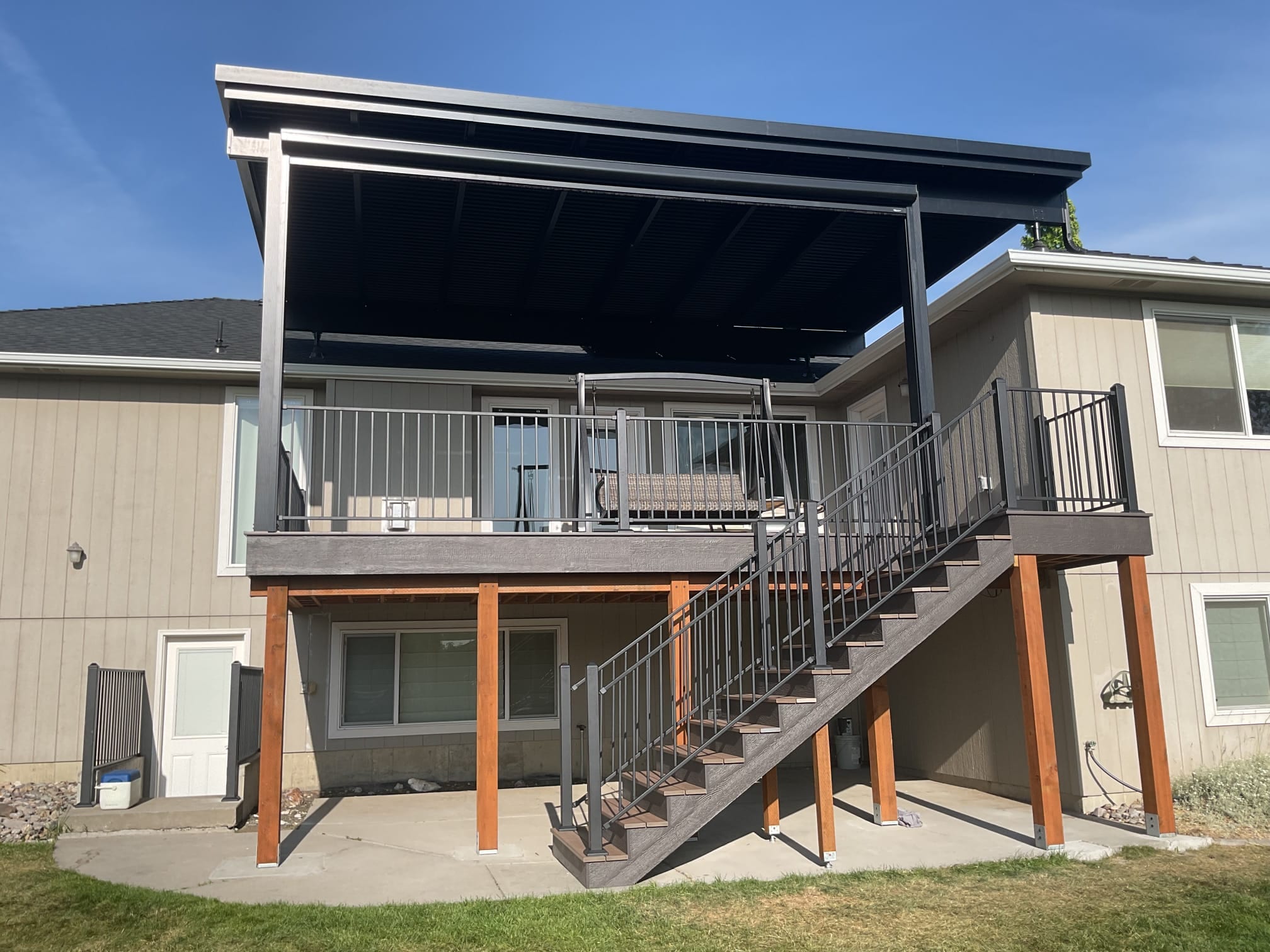 Elevated deck with dark flat louvered-style roof, gray metal railing and stairs on orange-wrapped posts over concrete patio