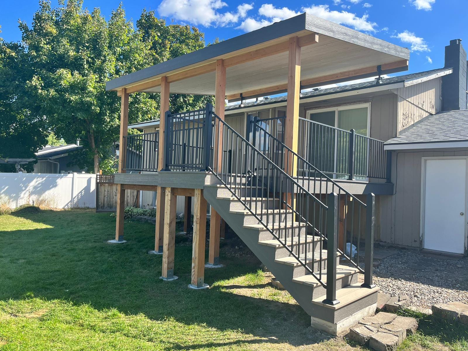 Elevated deck with flat metal roof cover on stained posts, black picket railing and wide stair, tan house and lawn