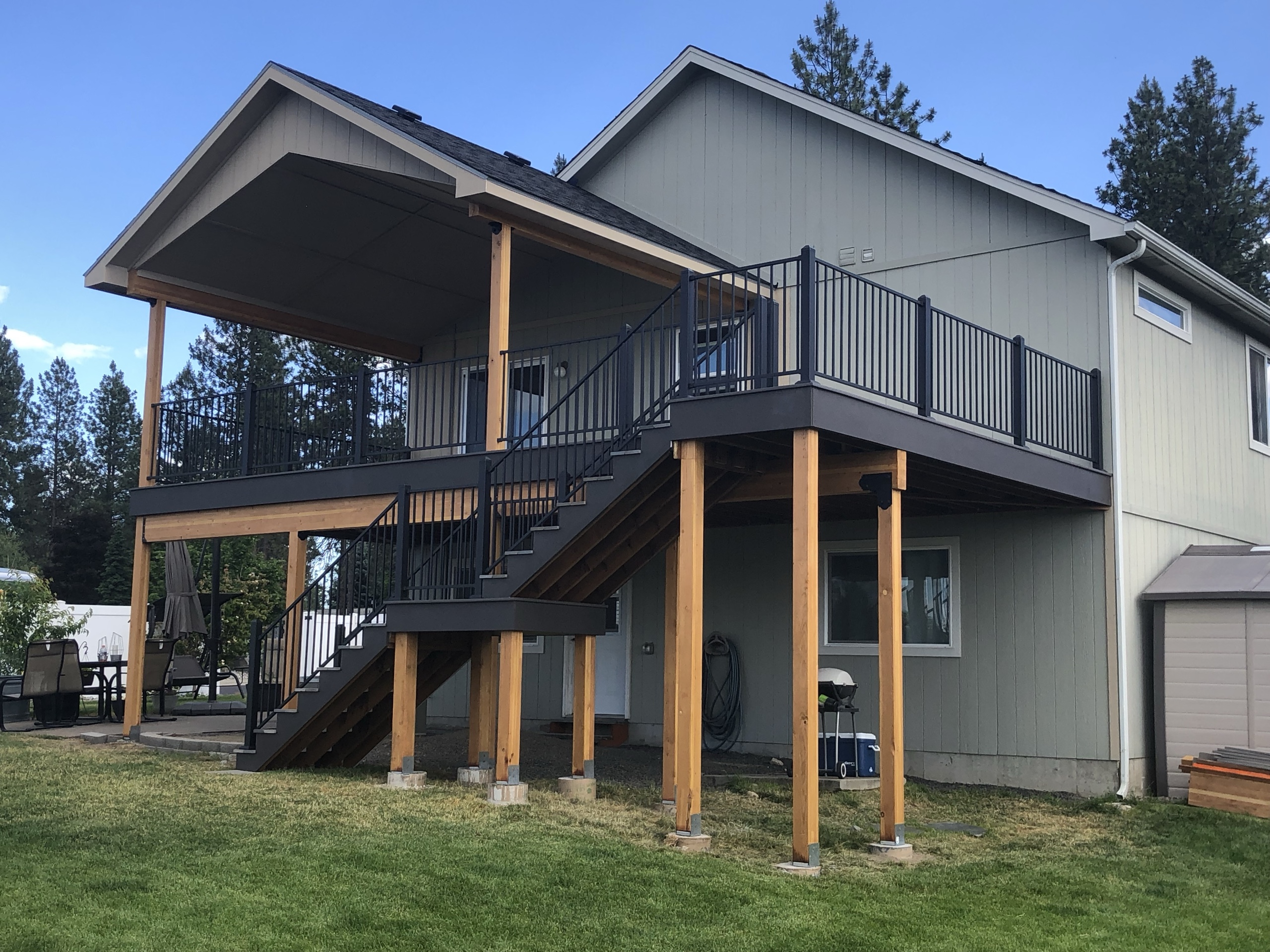 Elevated deck with gabled roof cover on cedar posts, black railing and switchback stairs on gray-green house