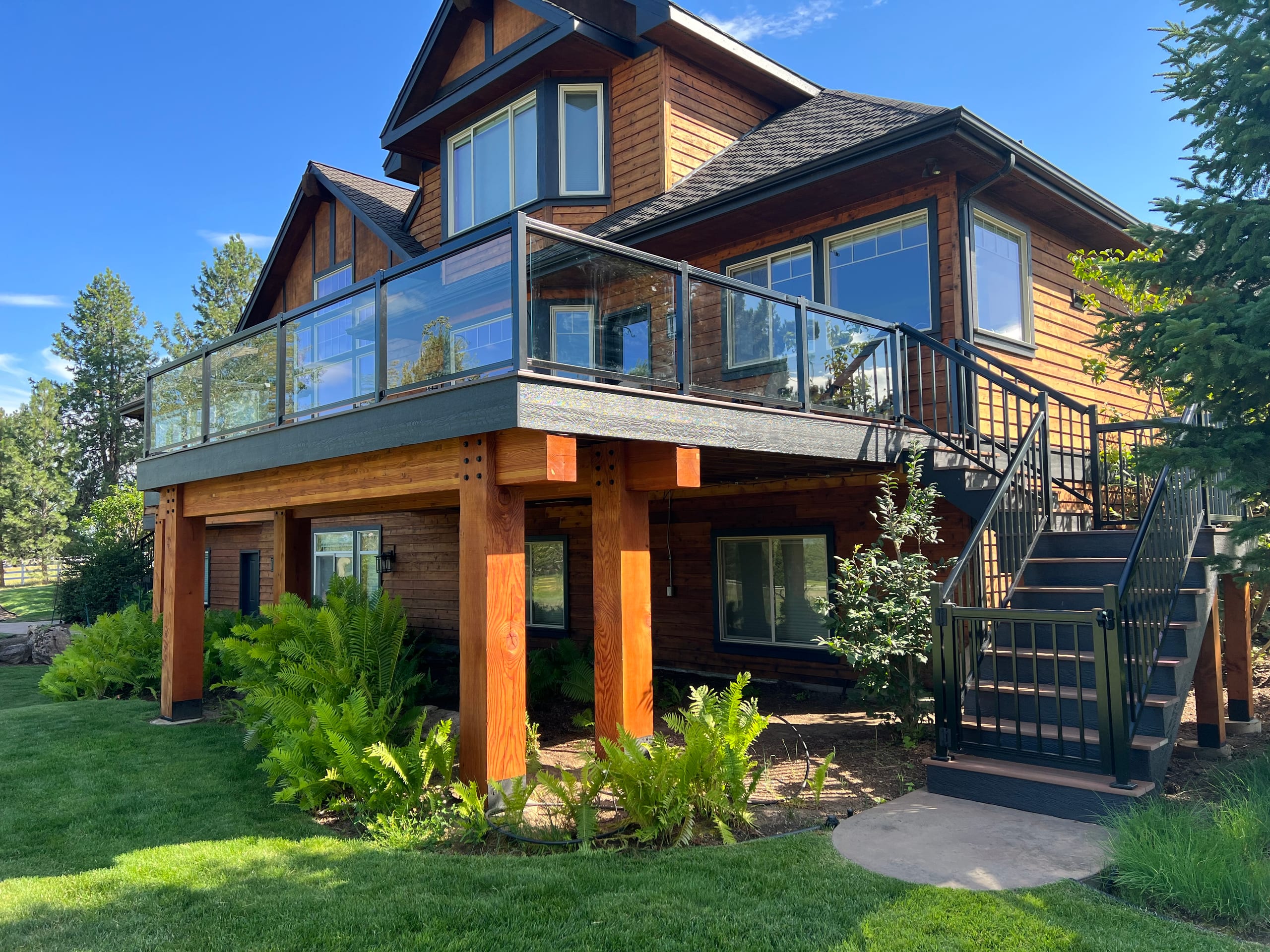 Elevated deck with glass railing on heavy timber posts beside a wood-sided house