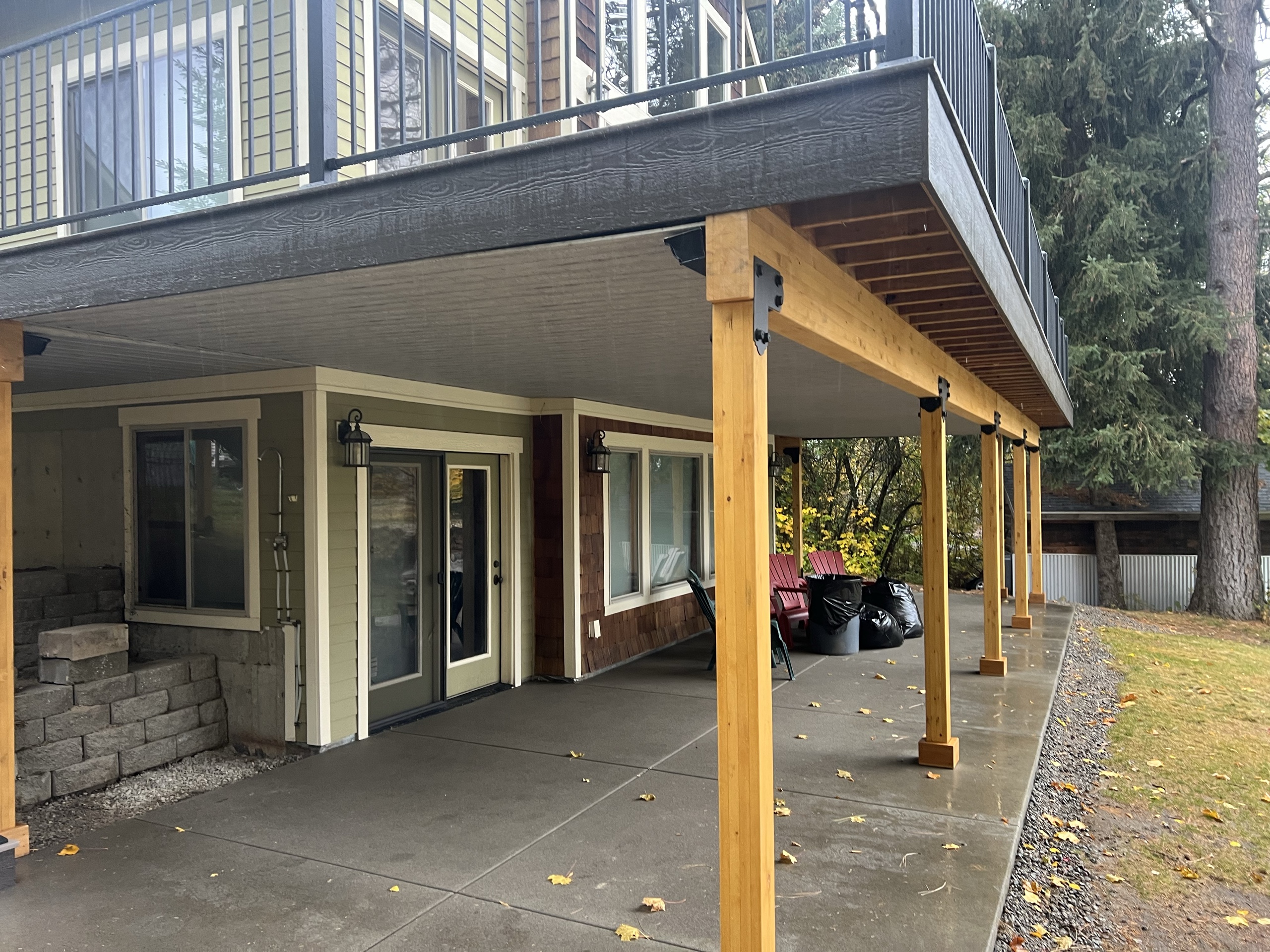 Elevated deck with glass-style railing above underdeck ceiling and cedar posts over a concrete patio, stone wall and doors