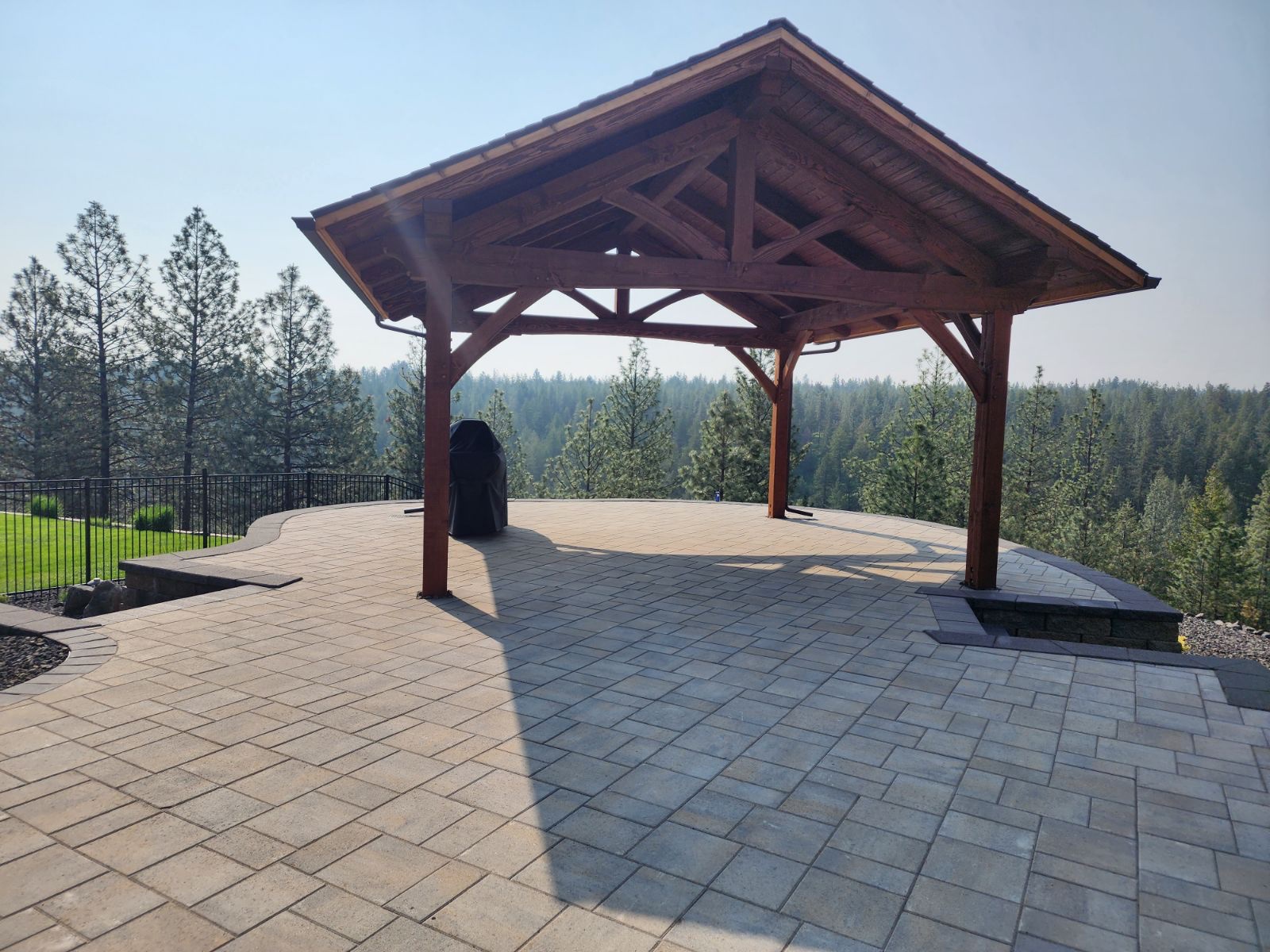 Freestanding timber gable pavilion on a paver patio overlooking pine forest, grill beneath, hazy sky
