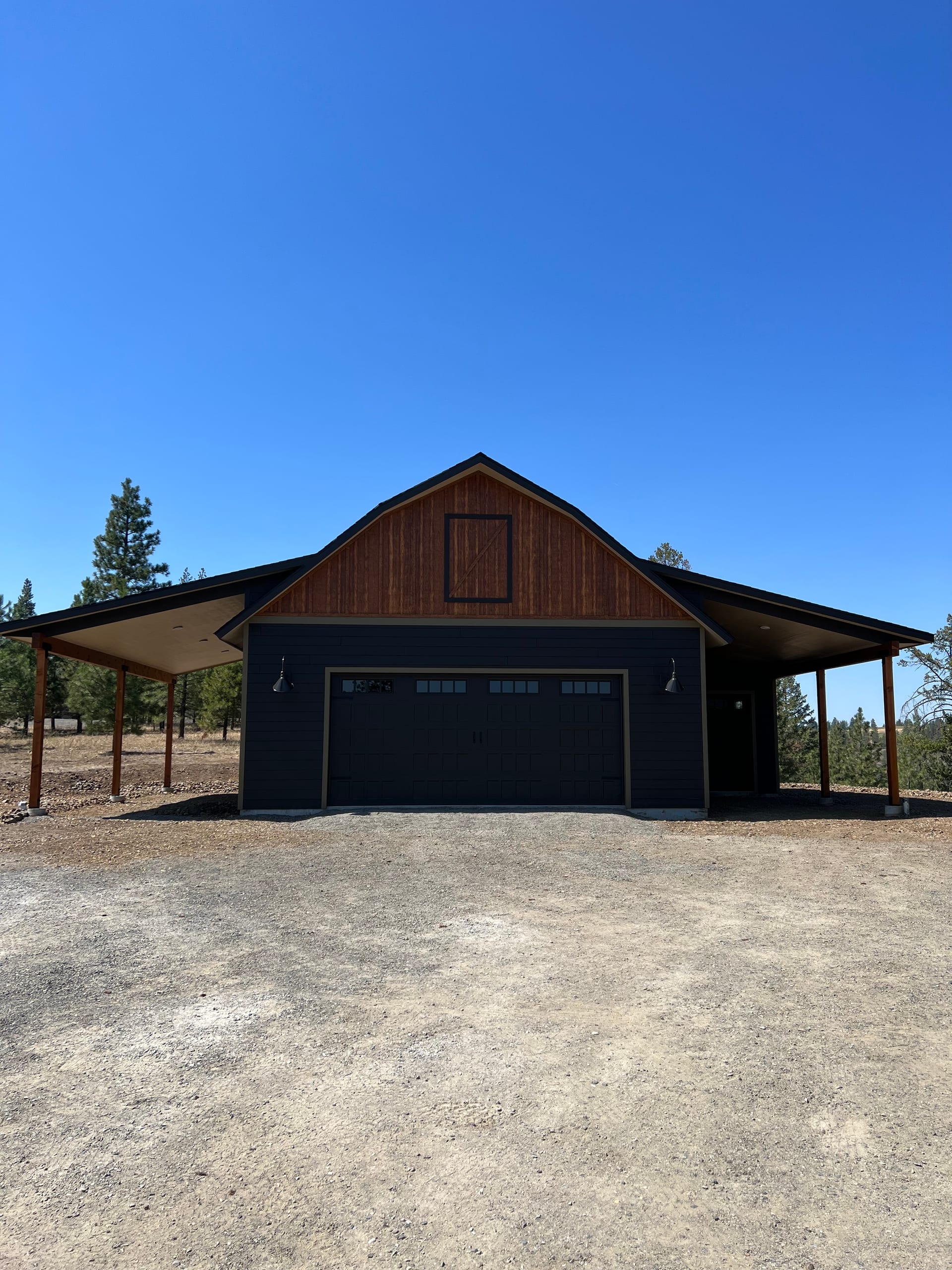 Front of dark barn-style garage with cedar gable and wrap-around covered carport on gravel, clear blue sky