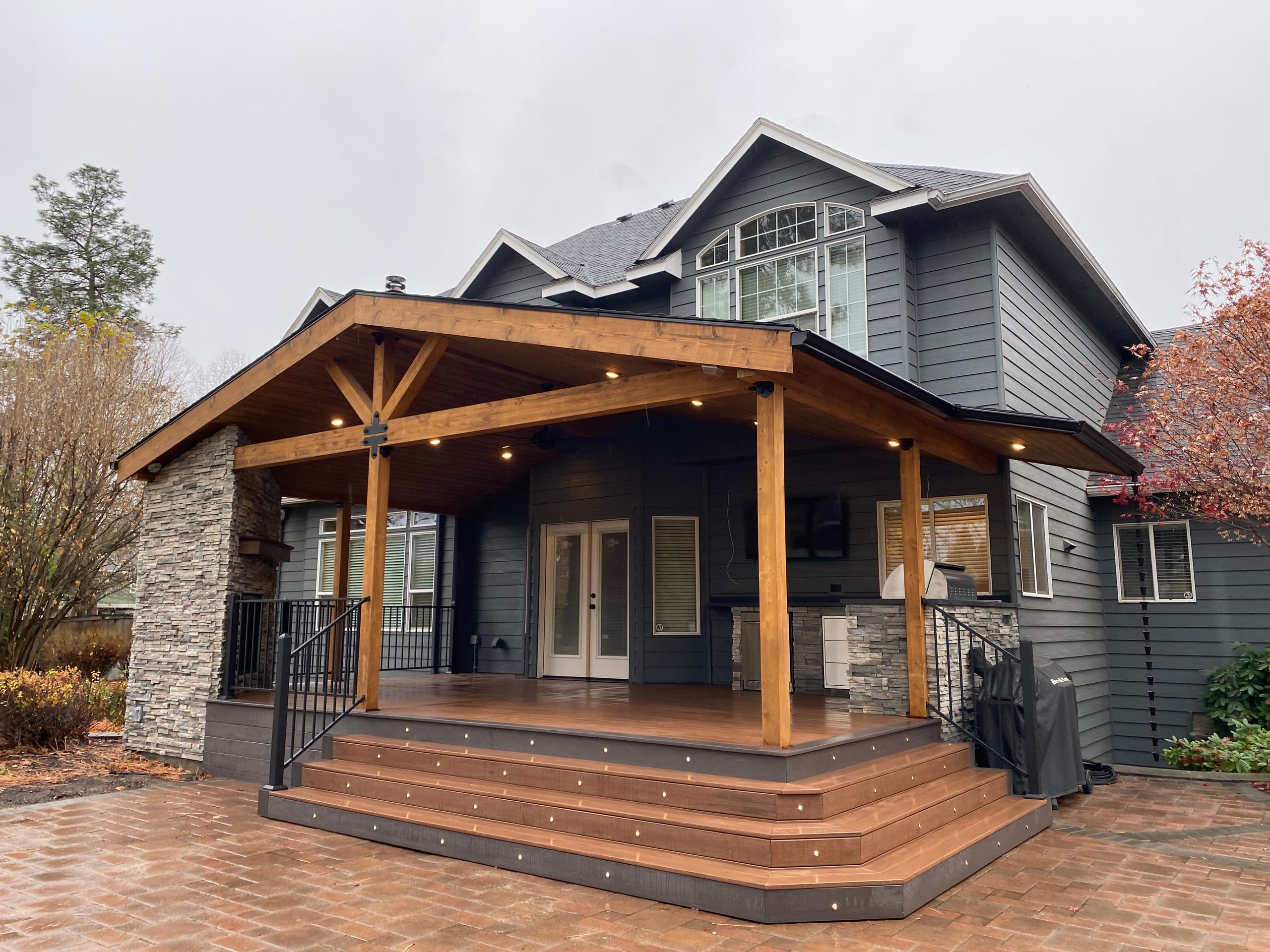Full gable covered deck with stone pier and wide step landing over a brick paver patio on a gray house, overcast