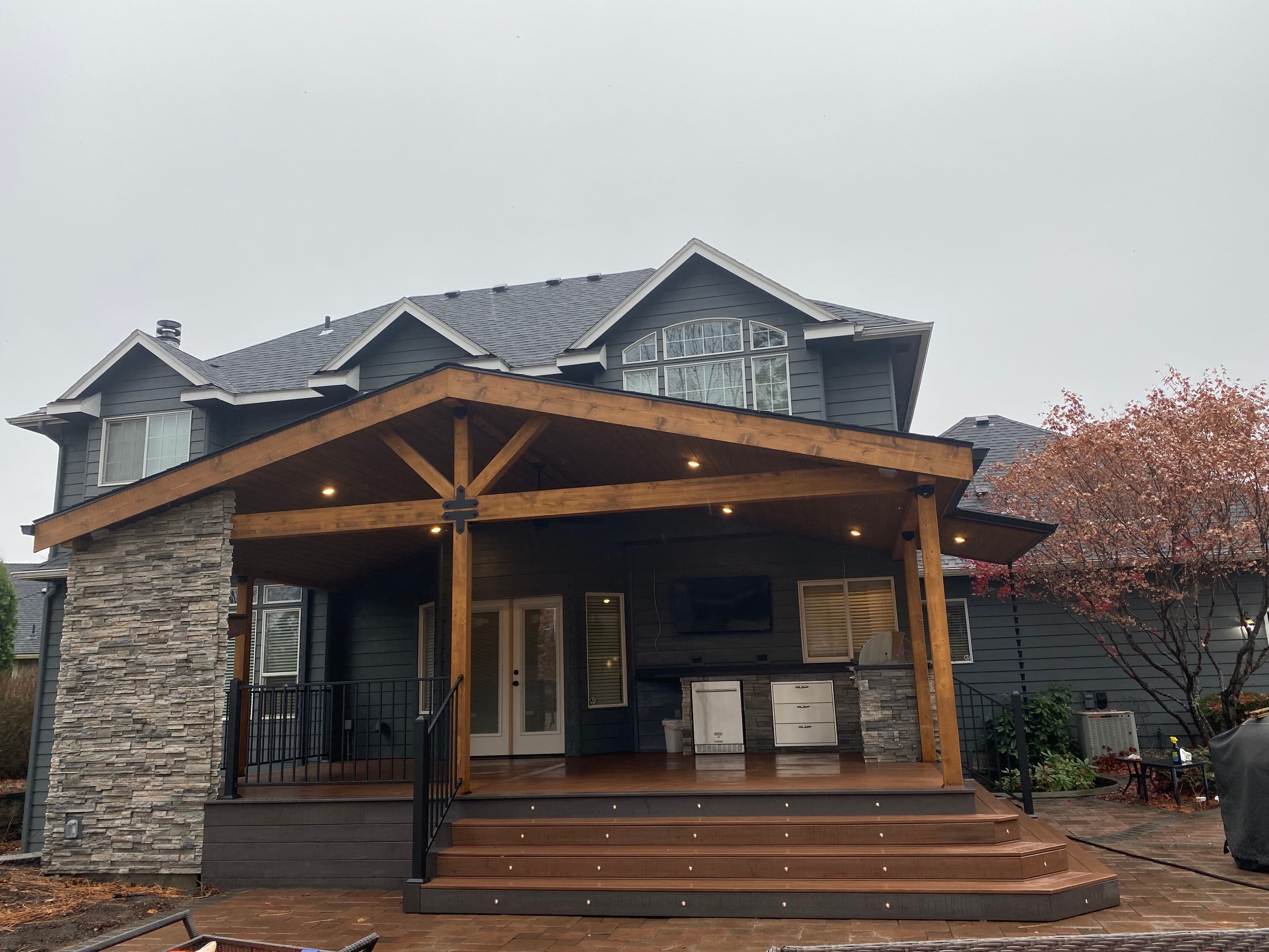 Gable covered deck with outdoor kitchen, stone pier and wide stepped landing on a paver patio, gray house, overcast