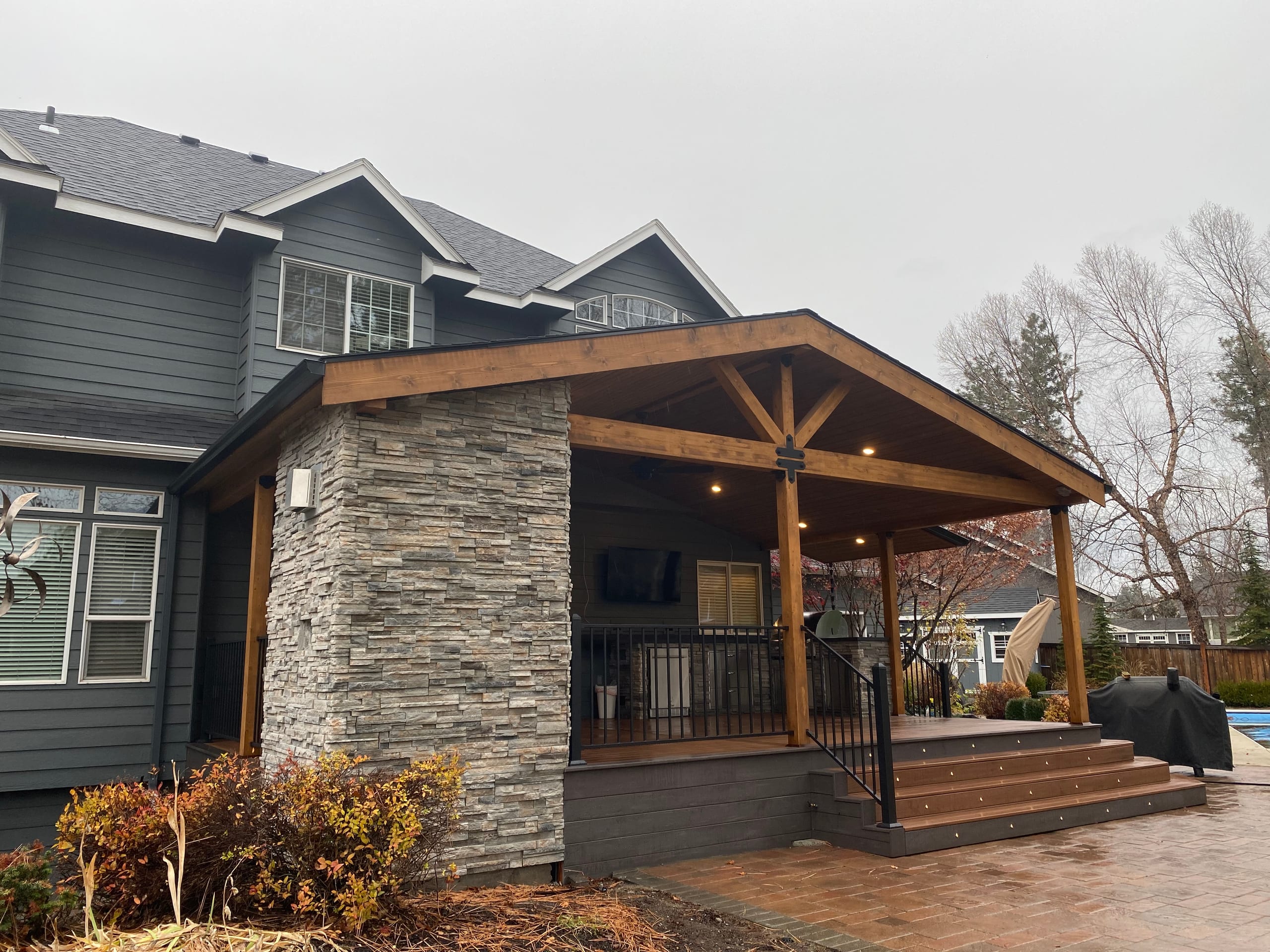 Gable covered deck with stained king-post truss, stone pillar, black railing and wide steps over a paver patio, overcast