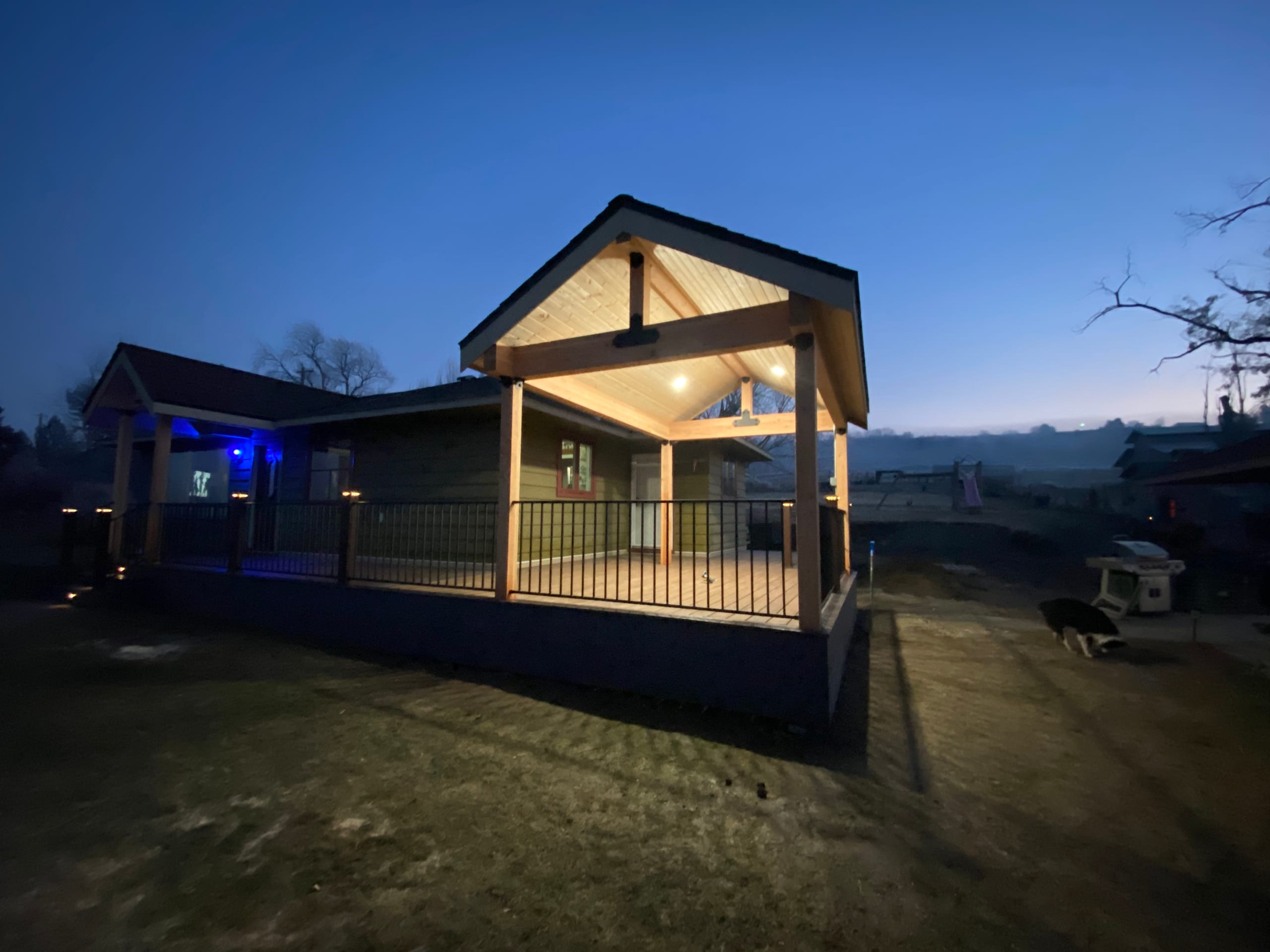 Gabled covered deck with glowing ceiling lights and black picket railing at blue hour, mountains behind