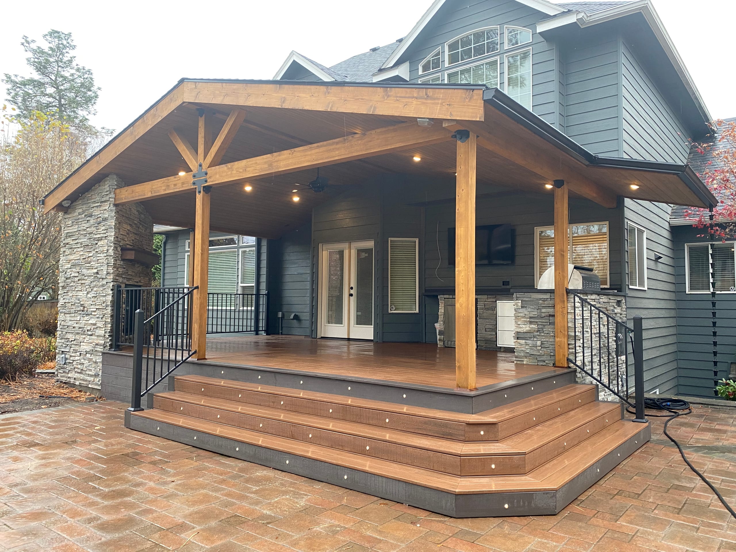 Gabled covered deck with stained timber trusses, stone pier, black railing