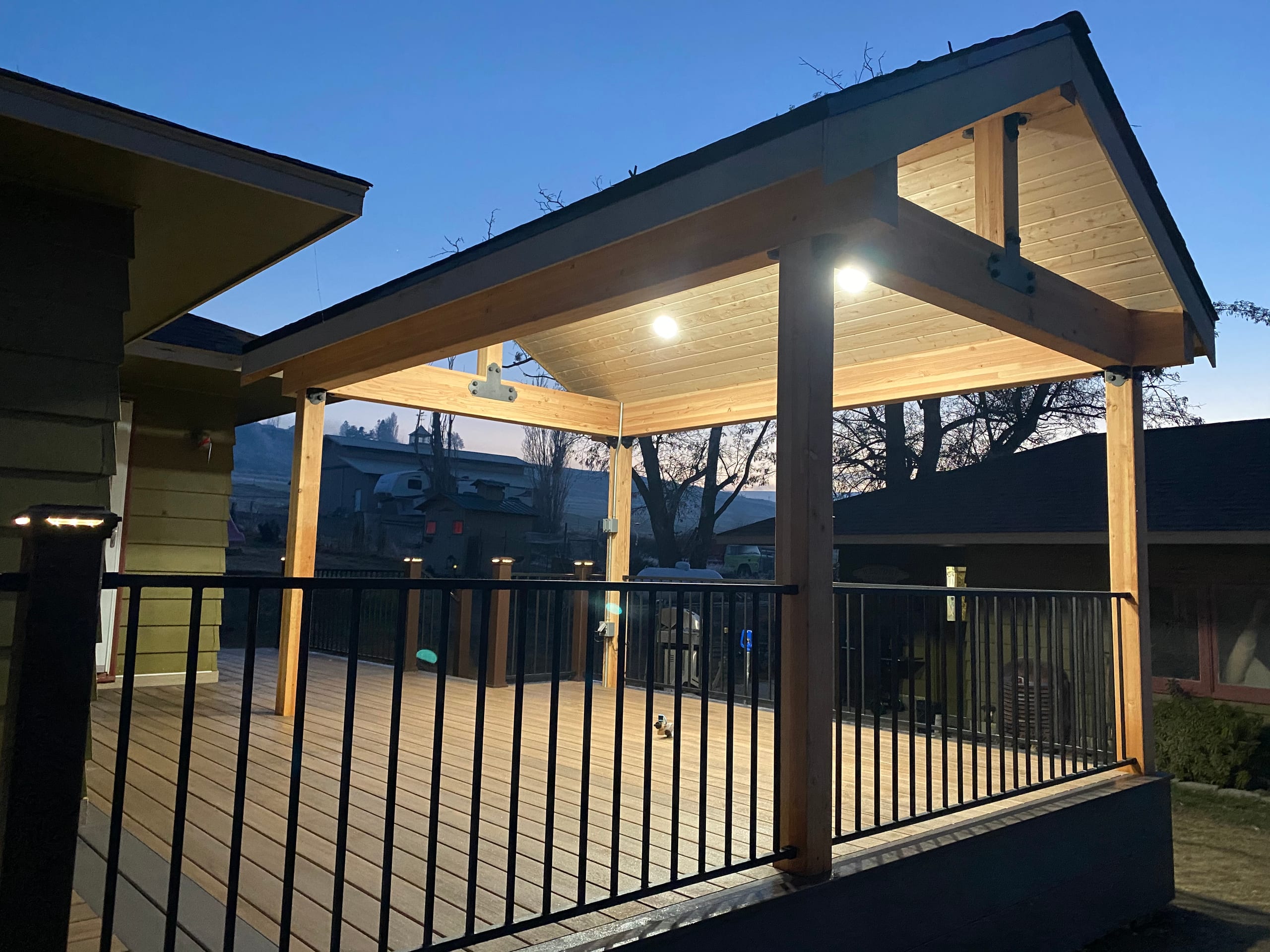 Gabled covered deck with timber posts, glowing recessed ceiling lights and black picket railing at dusk