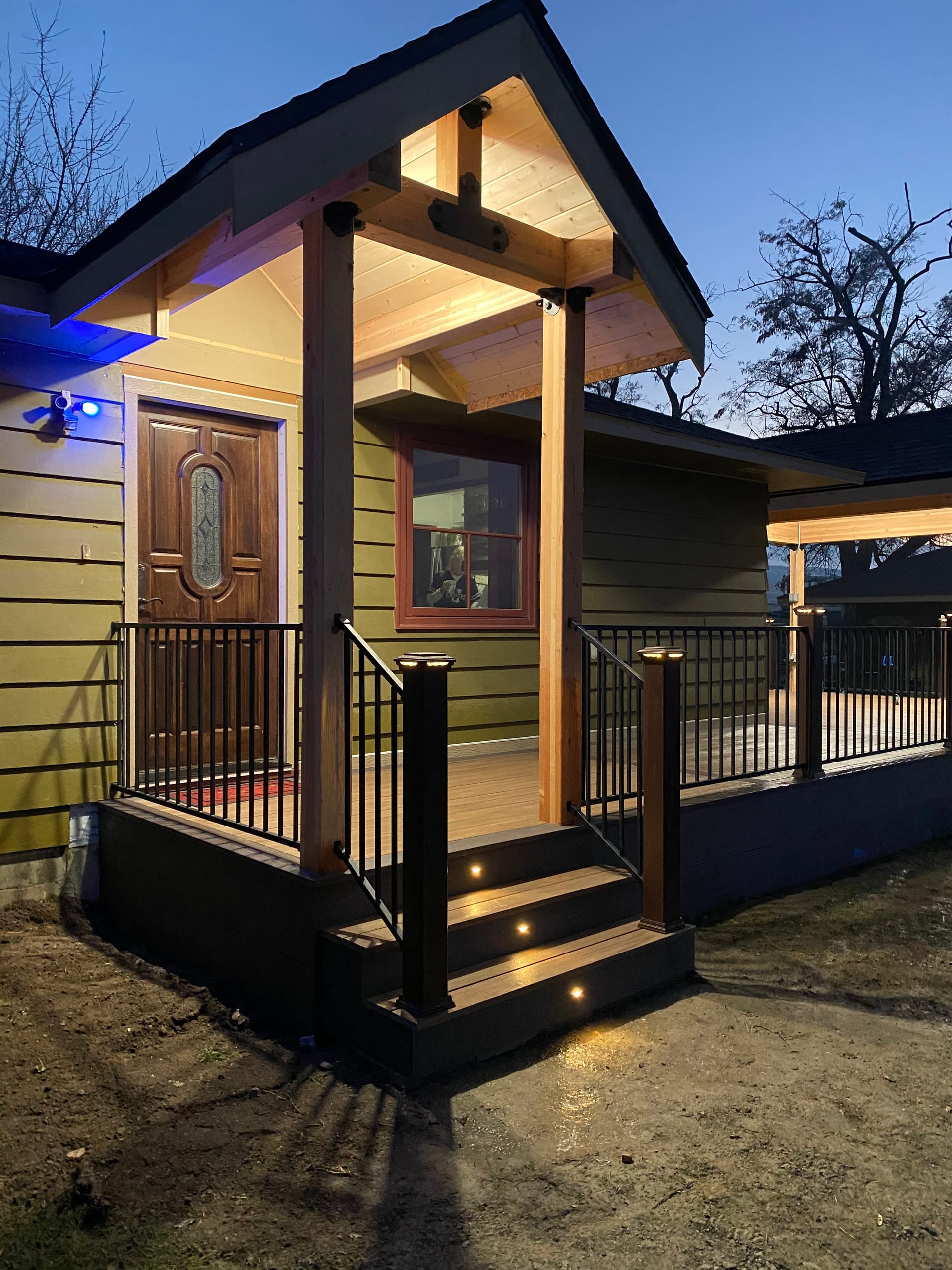 Gabled covered entry porch with glowing cedar ceiling, lit steps and black railing, adjacent covered deck behind, dusk