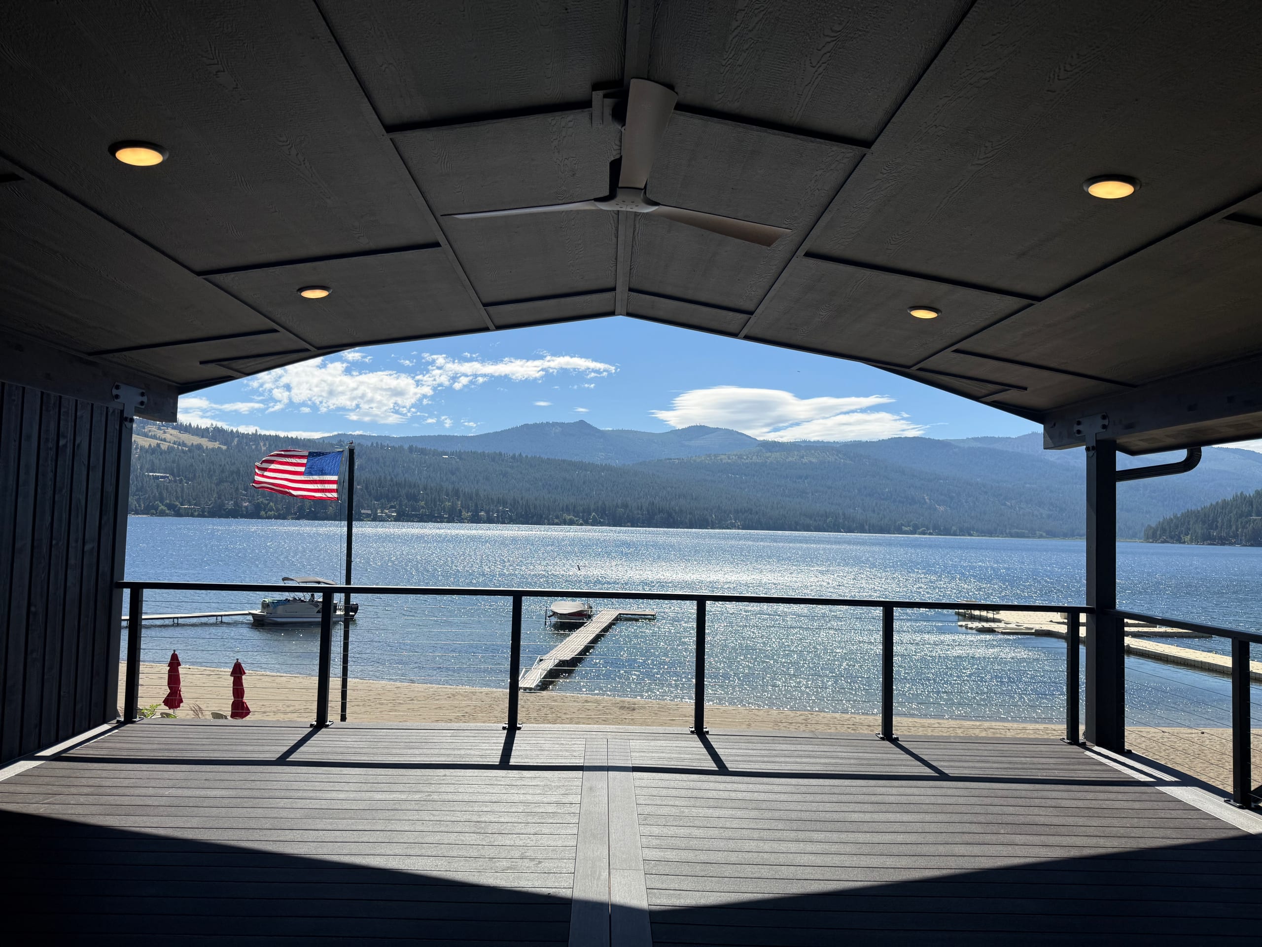 Gabled covered lakefront deck with lake, flag, dock and mountains seen through opening, fan and recessed lights