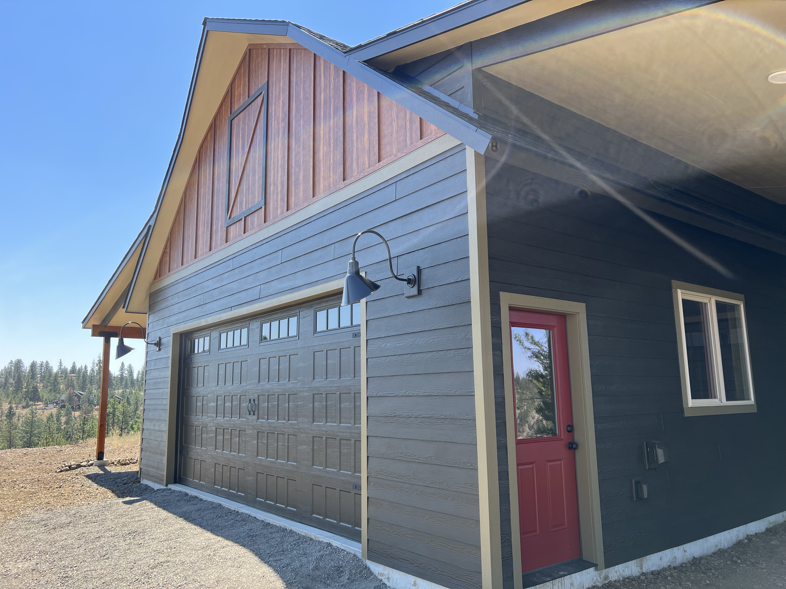 Corner of a dark-sided garage with a cedar gable, carriage-style garage door, red entry door and gooseneck light