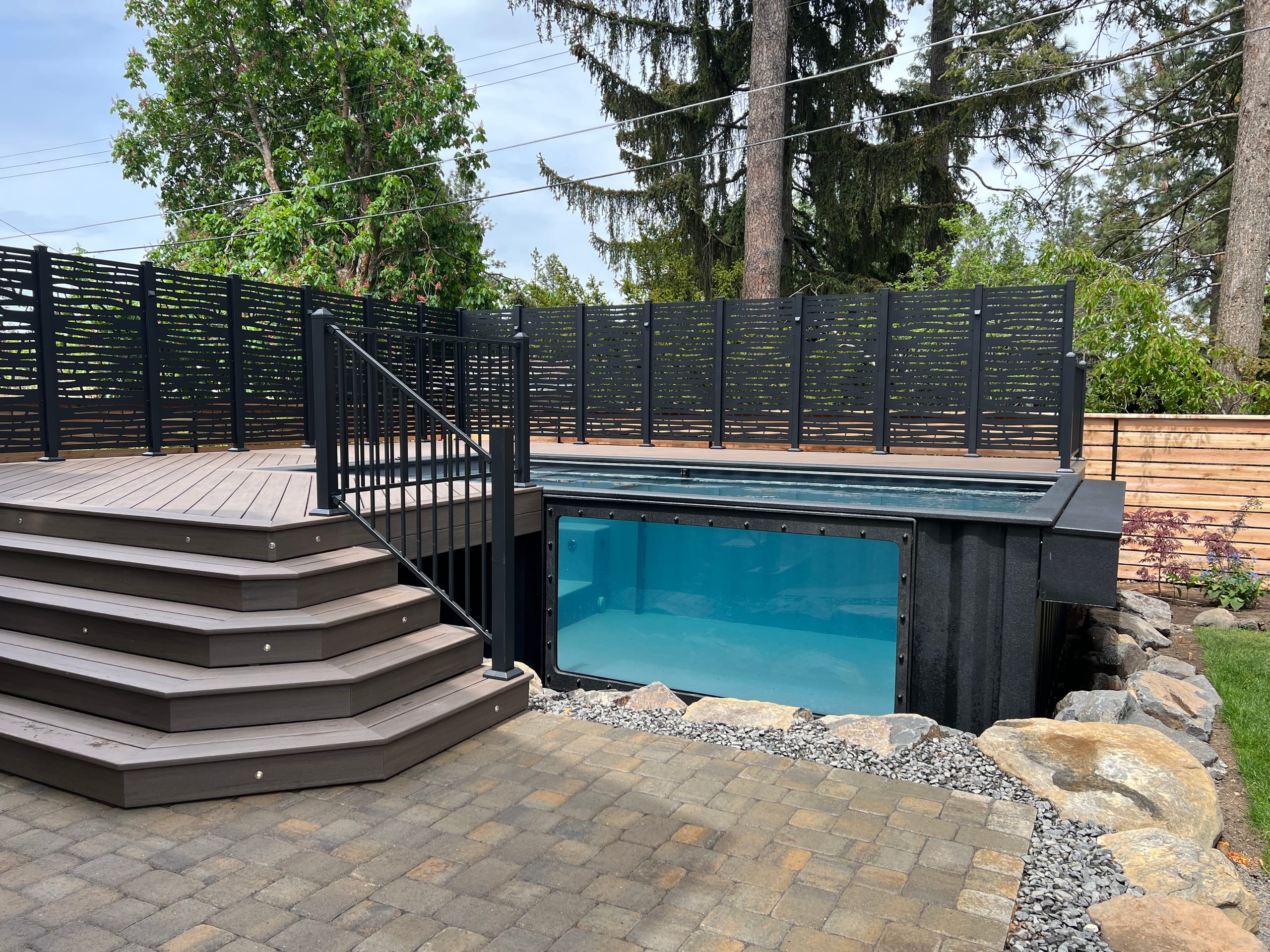 Glass-front raised pool in composite deck with corner steps, louvered black privacy screen and paver patio