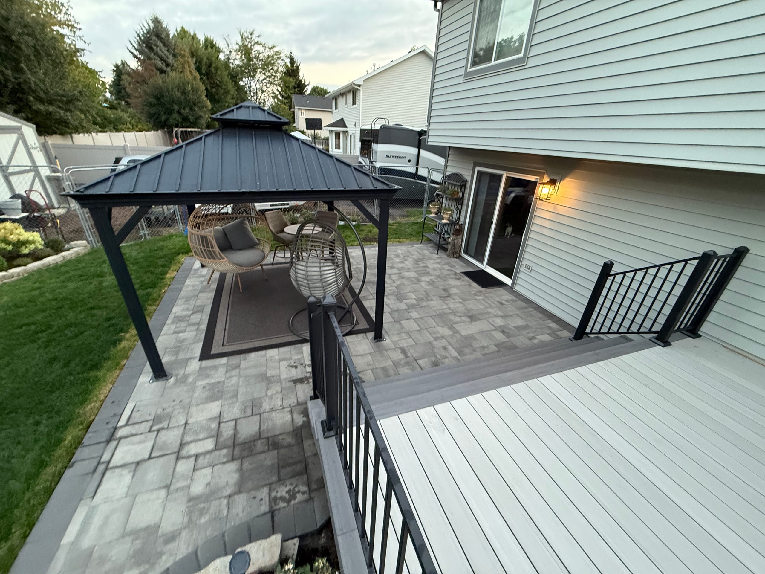 Gray composite deck with black picket railing stepping down to a paver patio and black gazebo with egg chairs, lawn beyond