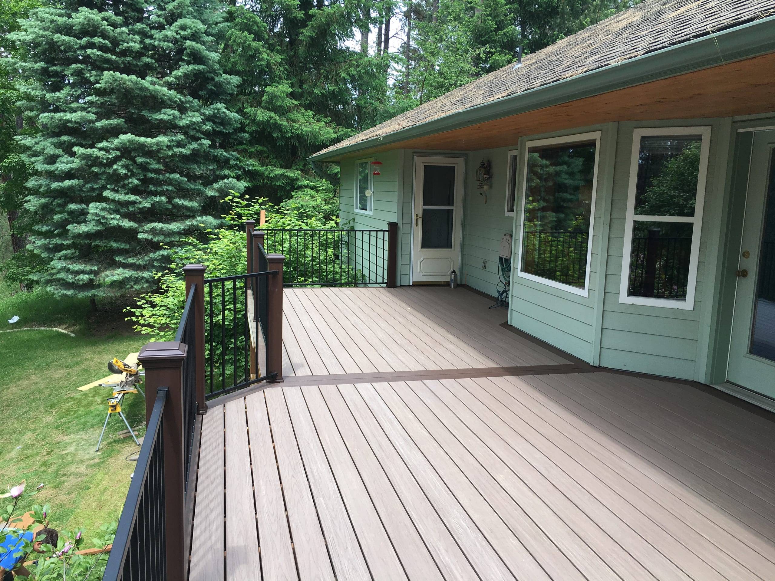 Gray composite deck with brown border inlay and black railing outside a sage-green house with cedar soffit, evergreens behind