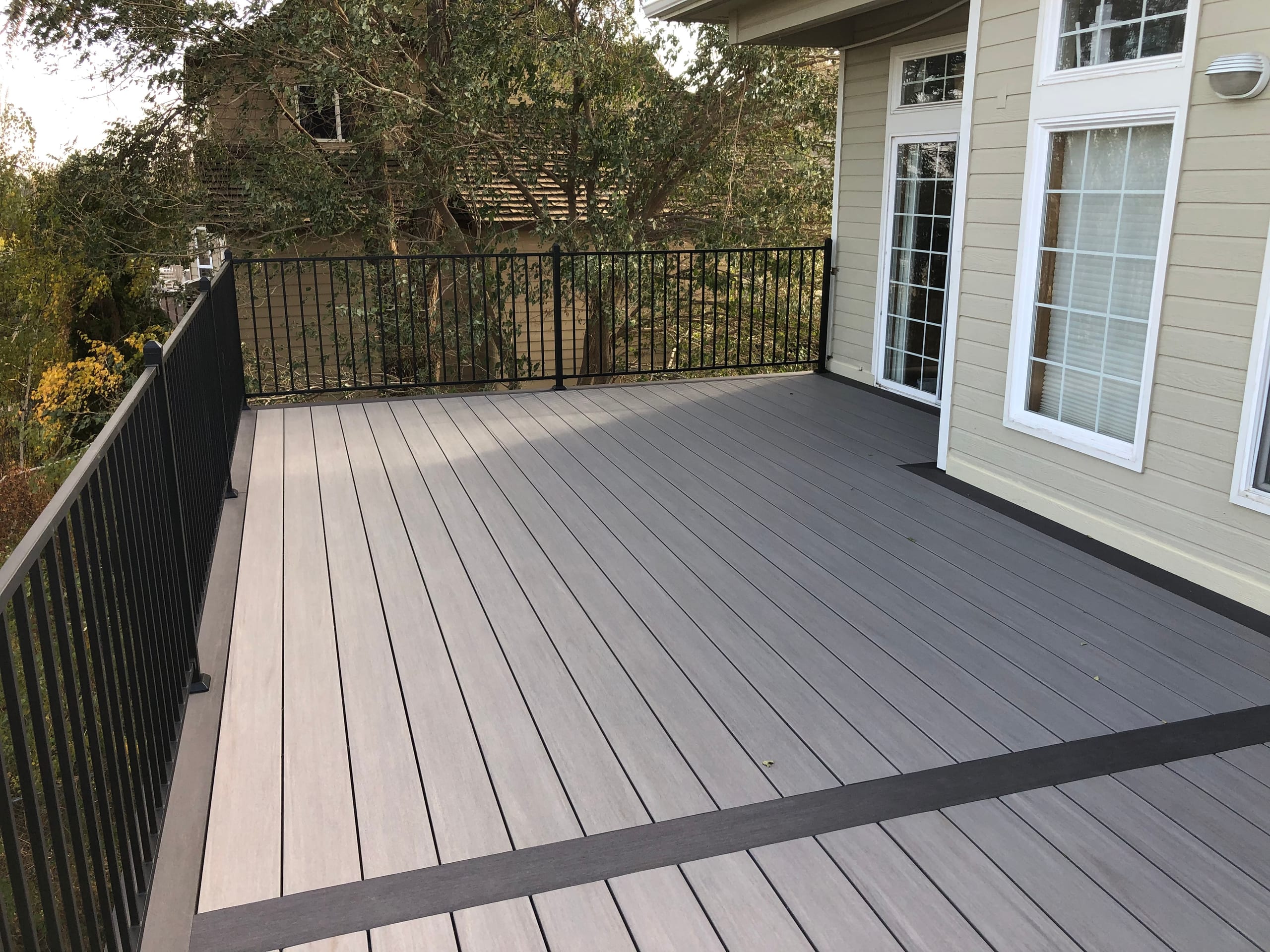 Gray composite deck with dark border and black metal picket railing beside beige house with tall windows, trees behind