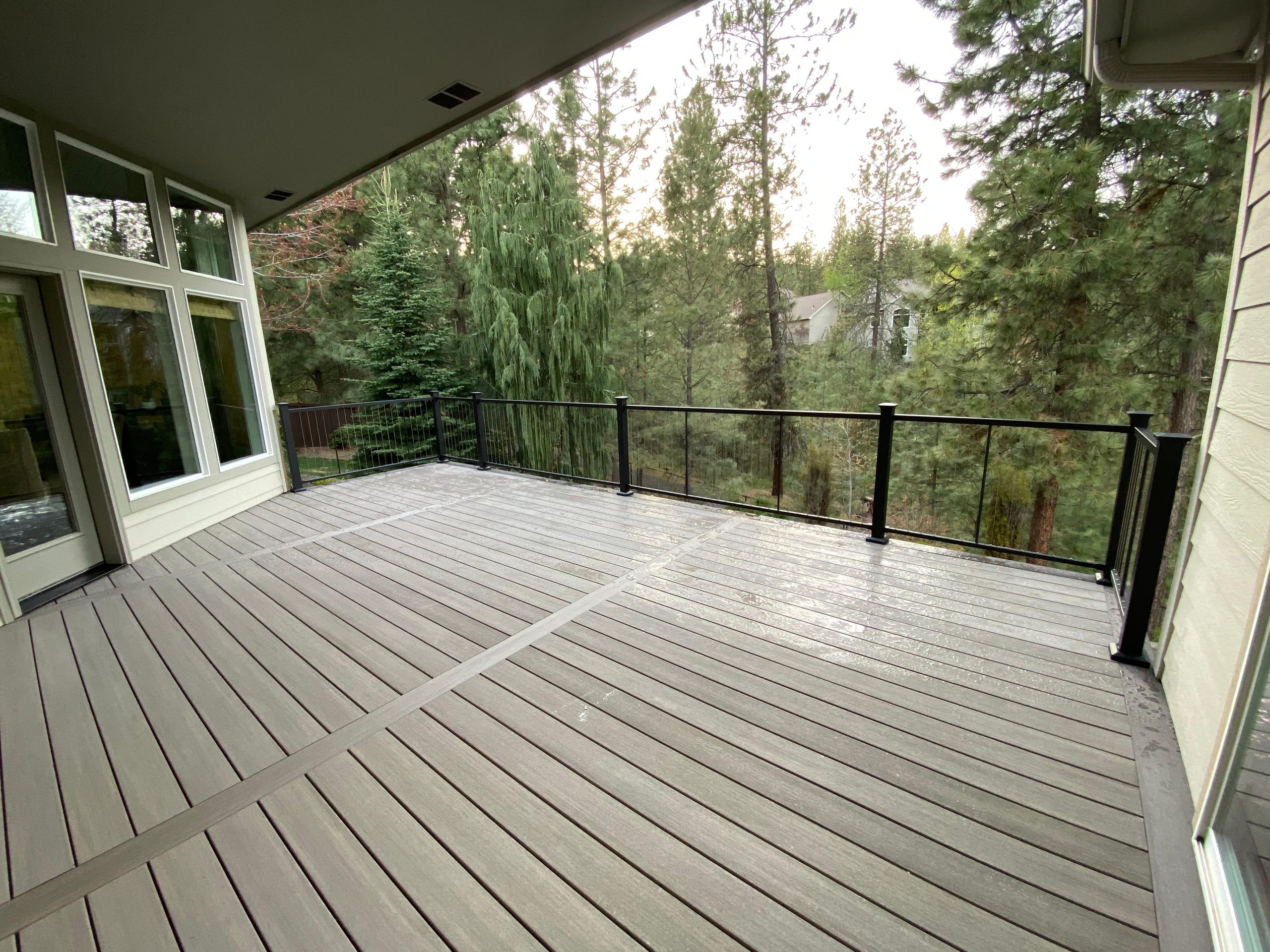 Gray composite deck with diagonal accent boards and black cable railing among pine trees, under roof overhang