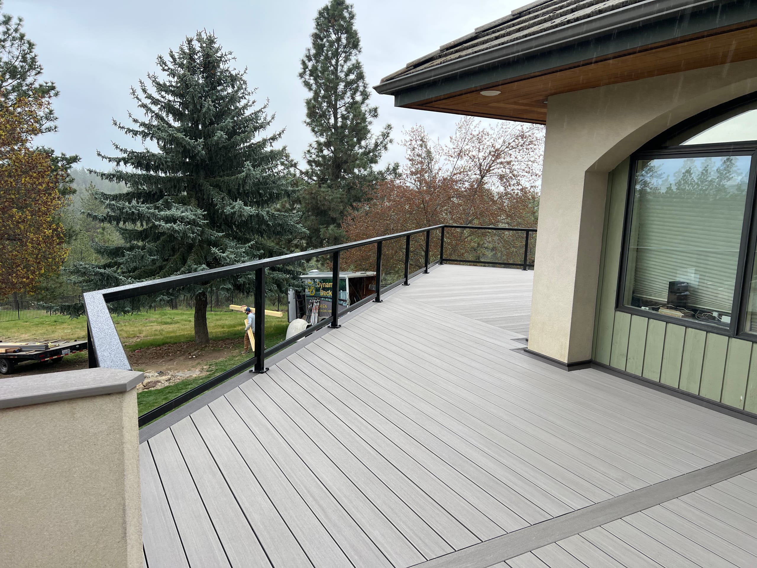 Second-story deck with gray decking and black-framed glass railing beside a stucco house