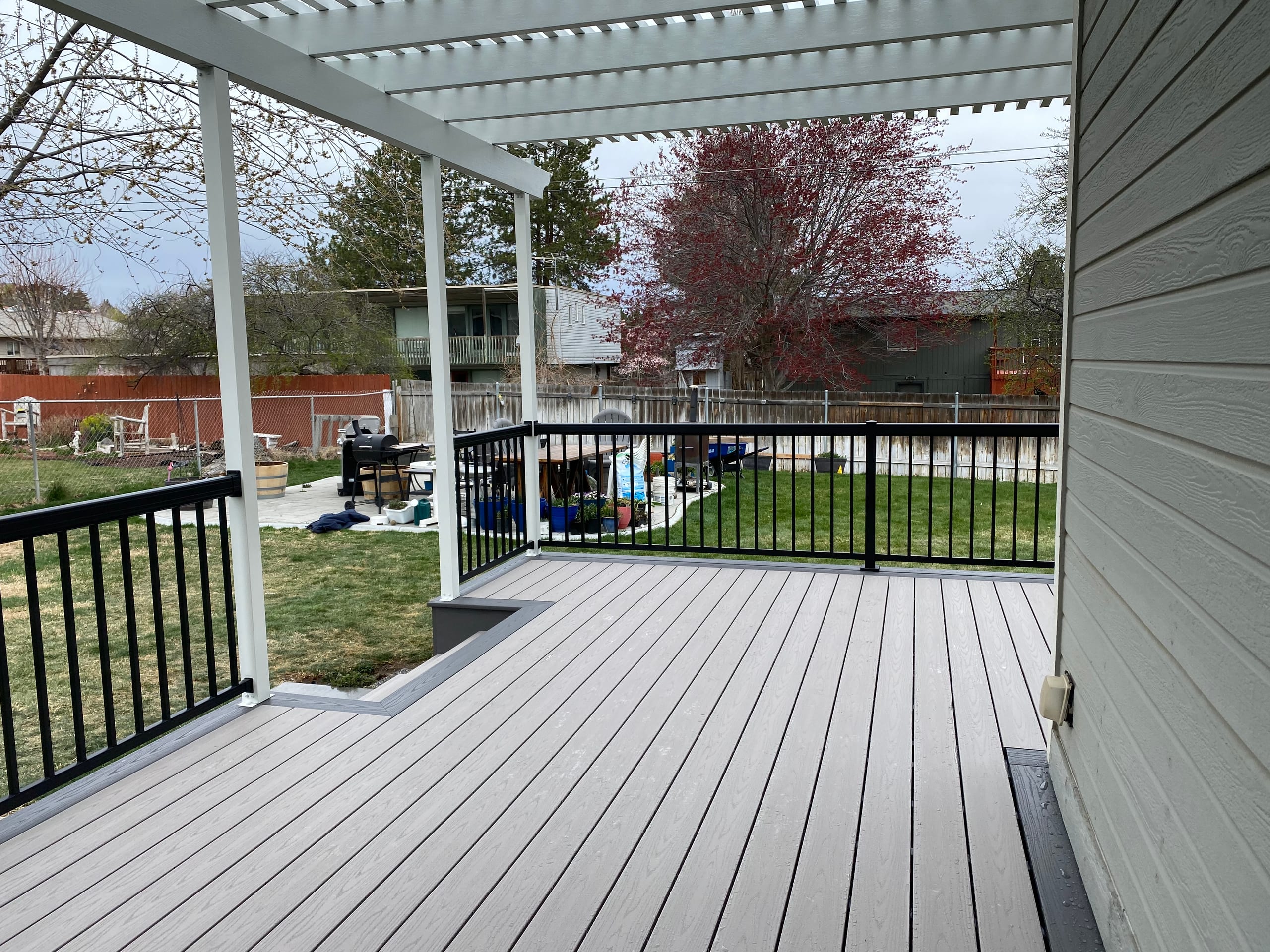 Gray deck partly under a white pergola with black railing overlooking a fenced backyard