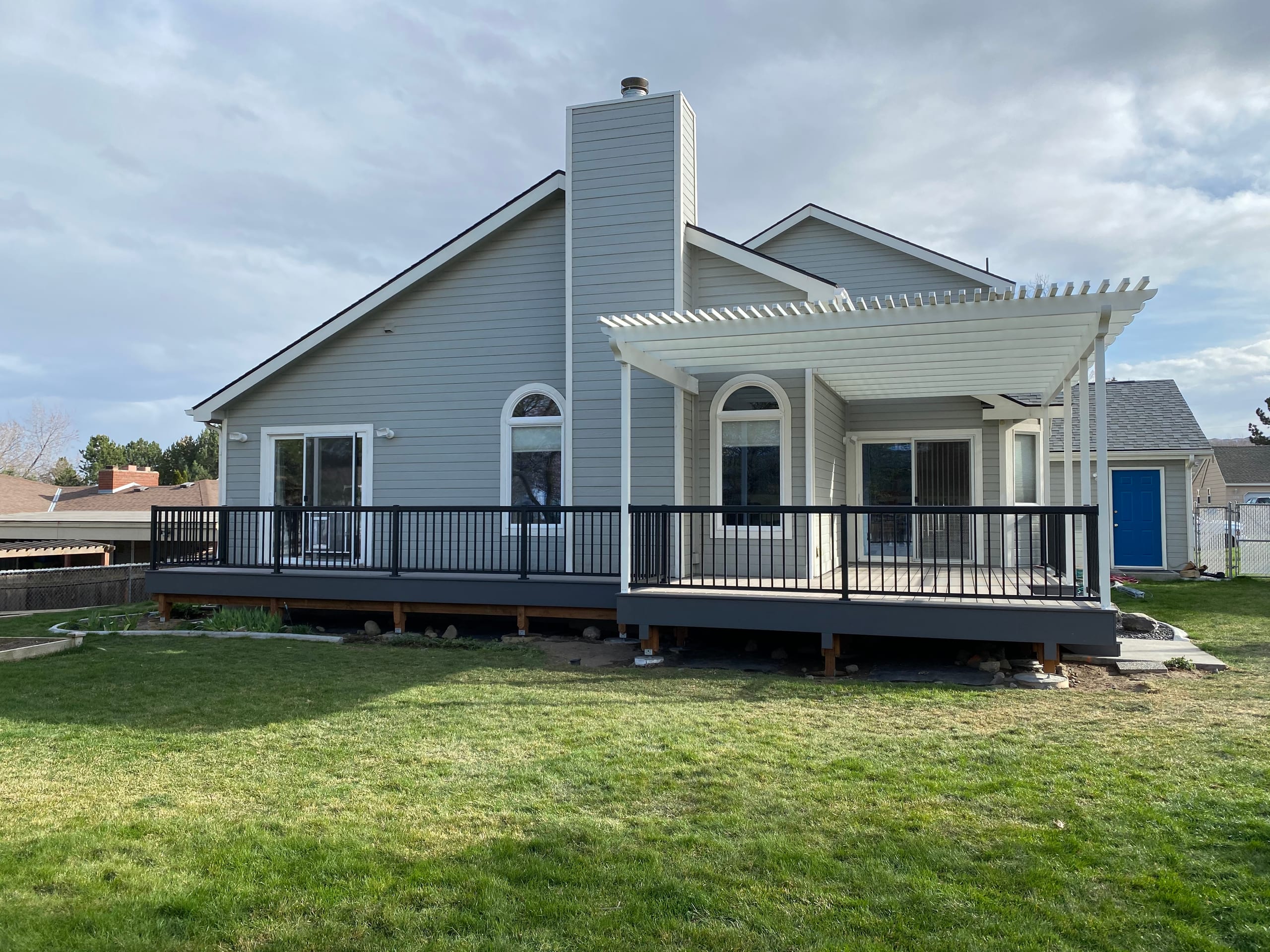 Gray house back with long dark-railed gray deck and a white louvered pergola over one end, lawn