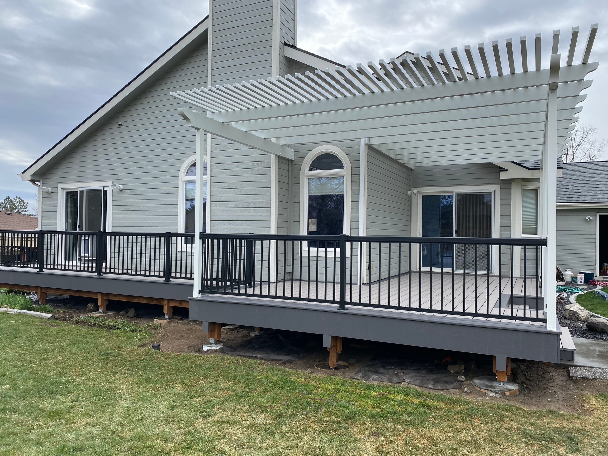 Gray house with light gray composite deck, white louvered aluminum pergola, black picket railing and dark skirt, lawn