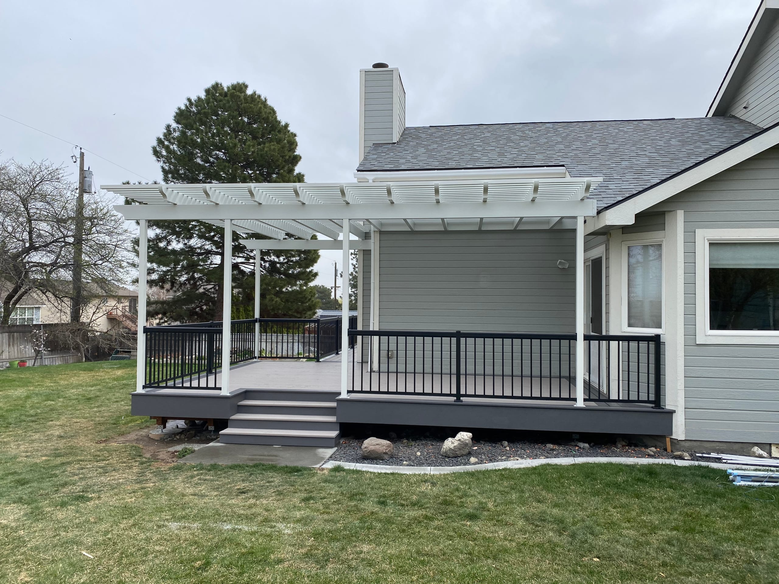 Gray house with white louvered pergola over gray composite deck with black railing and steps, lawn