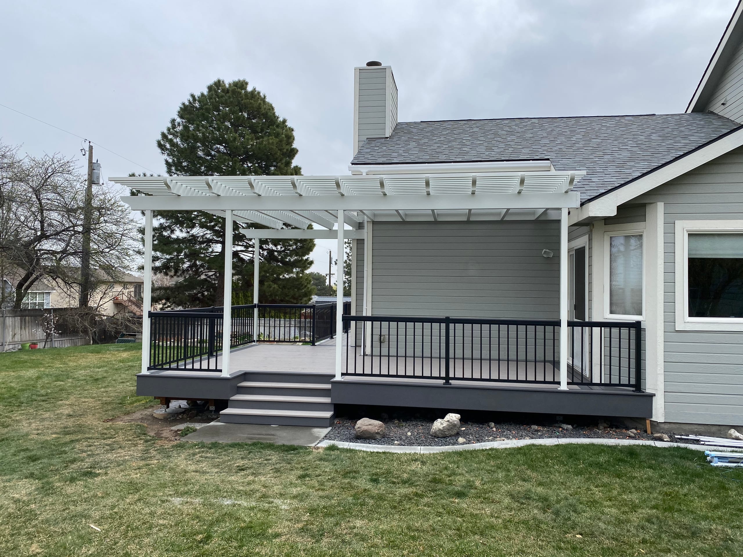 Gray house with white louvered pergola on posts over gray composite deck with black railing, steps and rock border