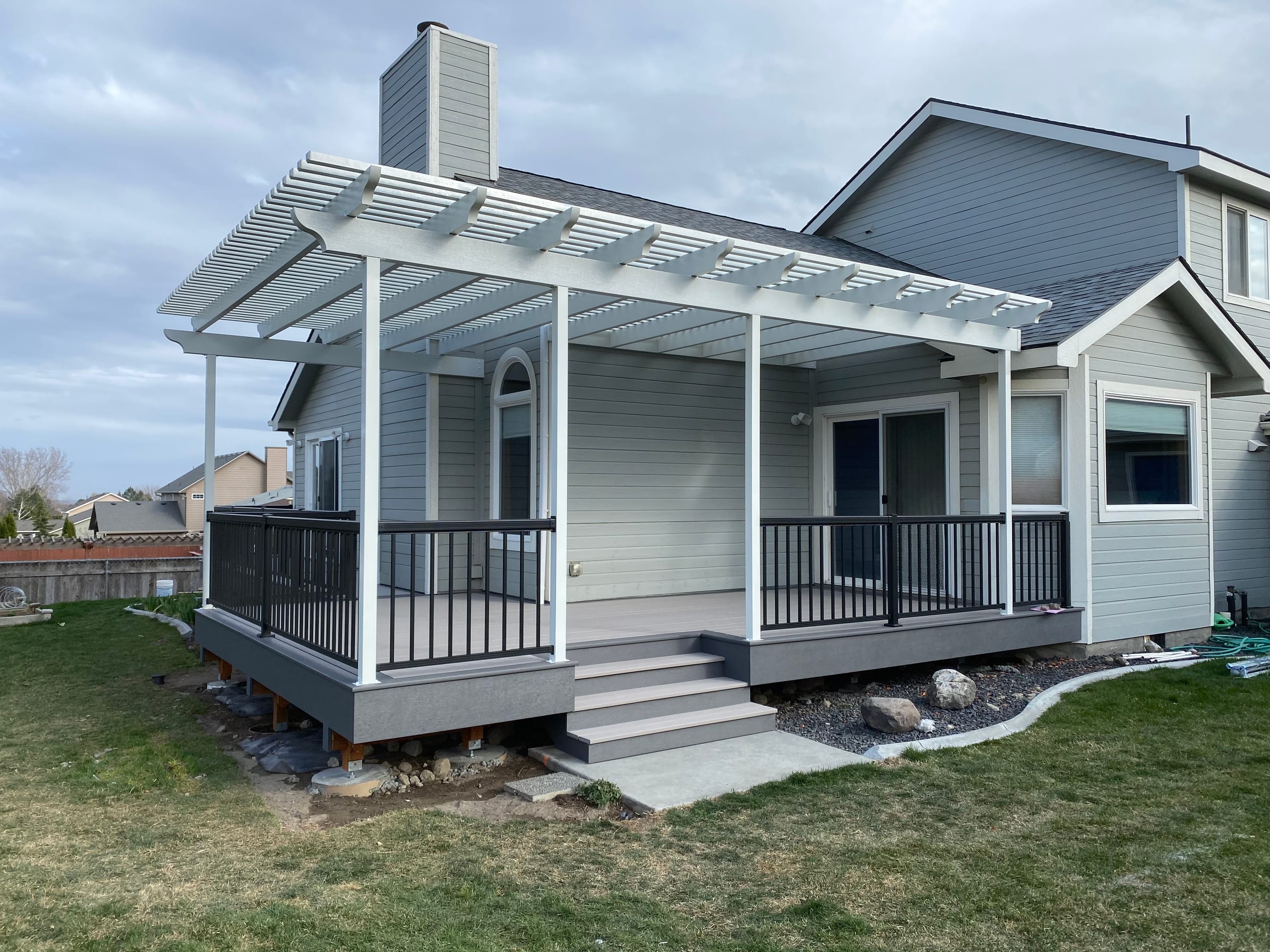Gray house with white pergola, gray composite deck with black railing and steps, landscaped edge