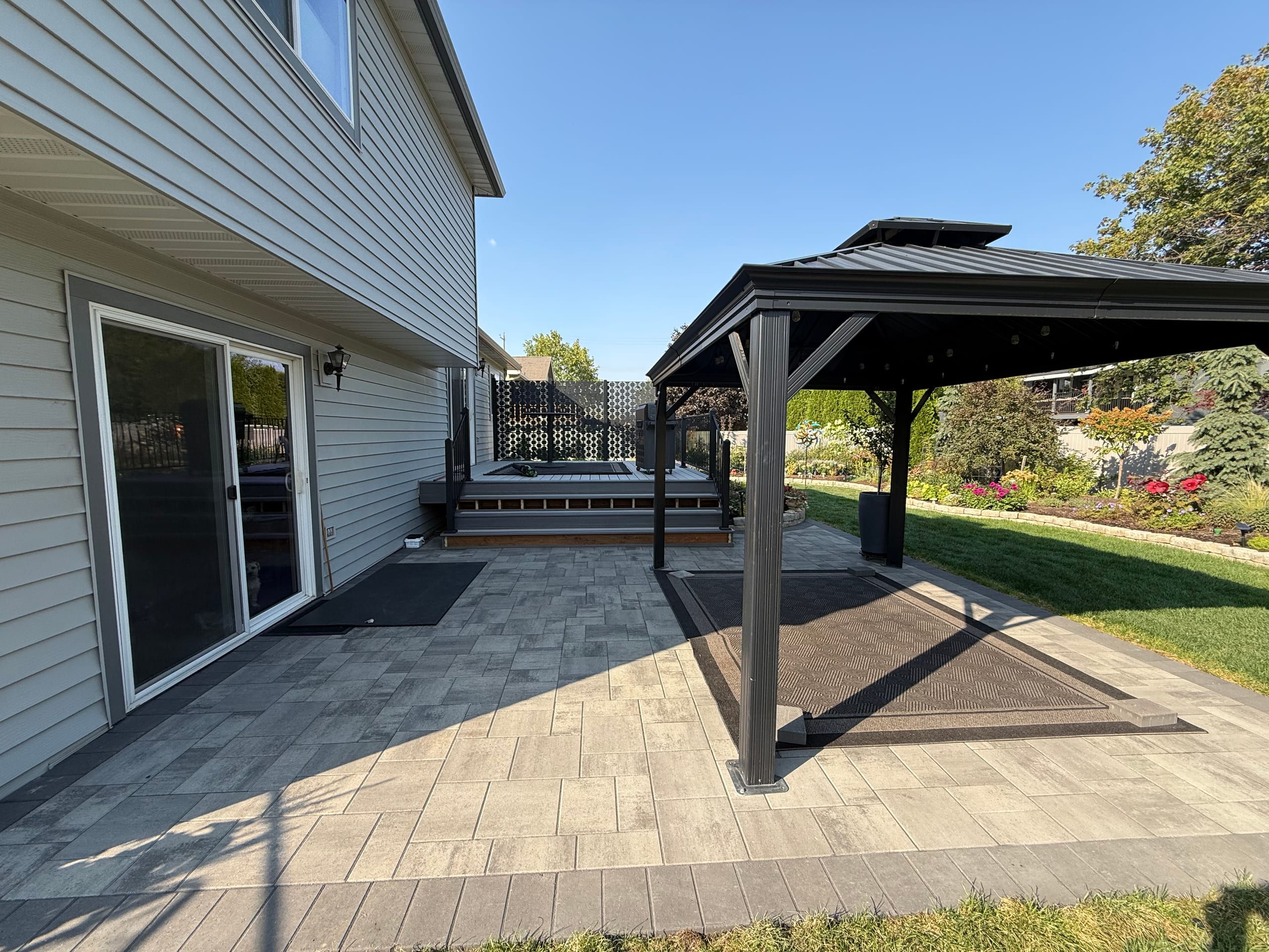 Gray paver patio with black metal hardtop gazebo, rug and seating, beside a gray two-story house, blue sky