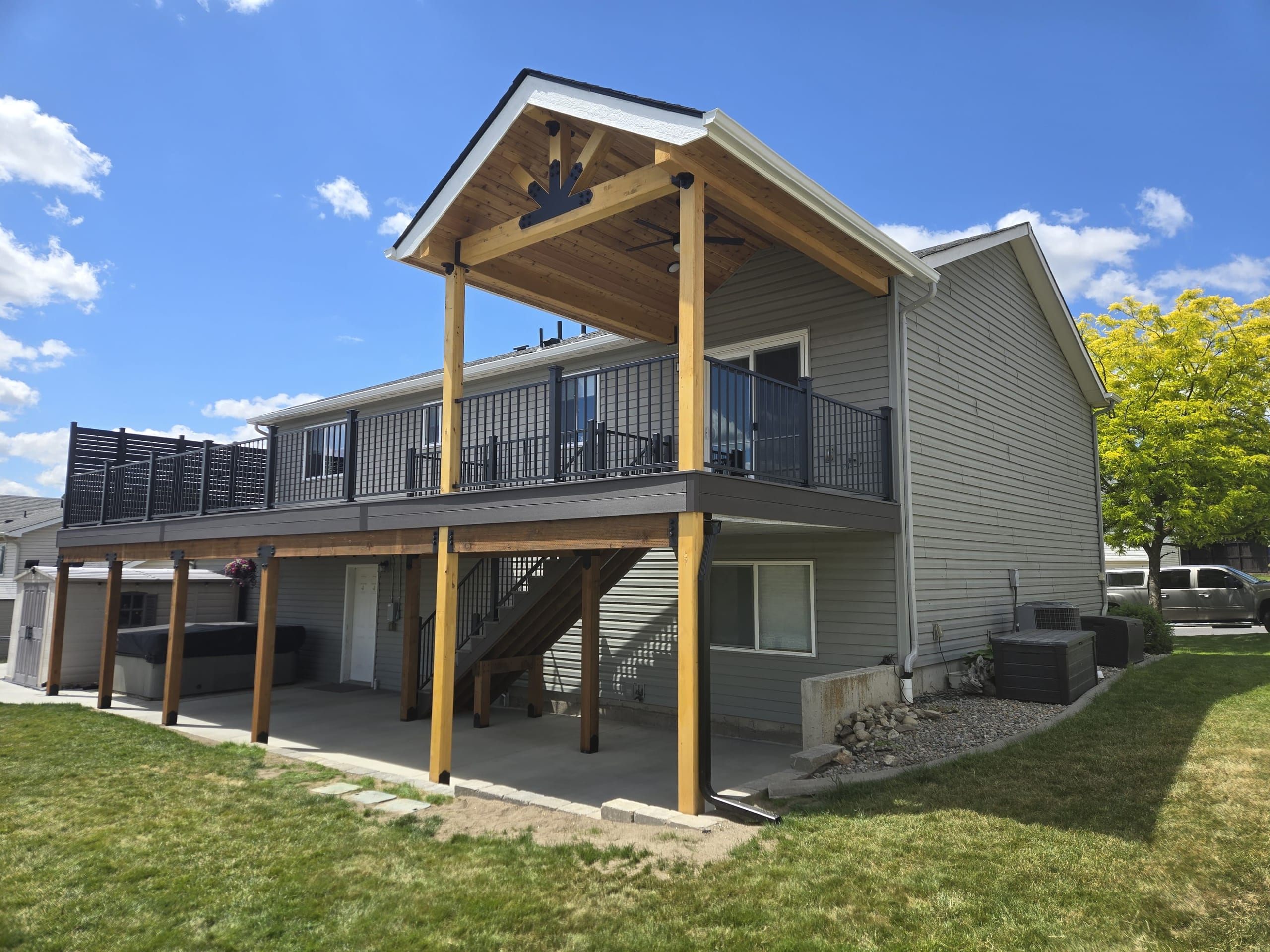 Gray two-story home with elevated deck with black railing and gabled cedar-post cover over covered patio beneath