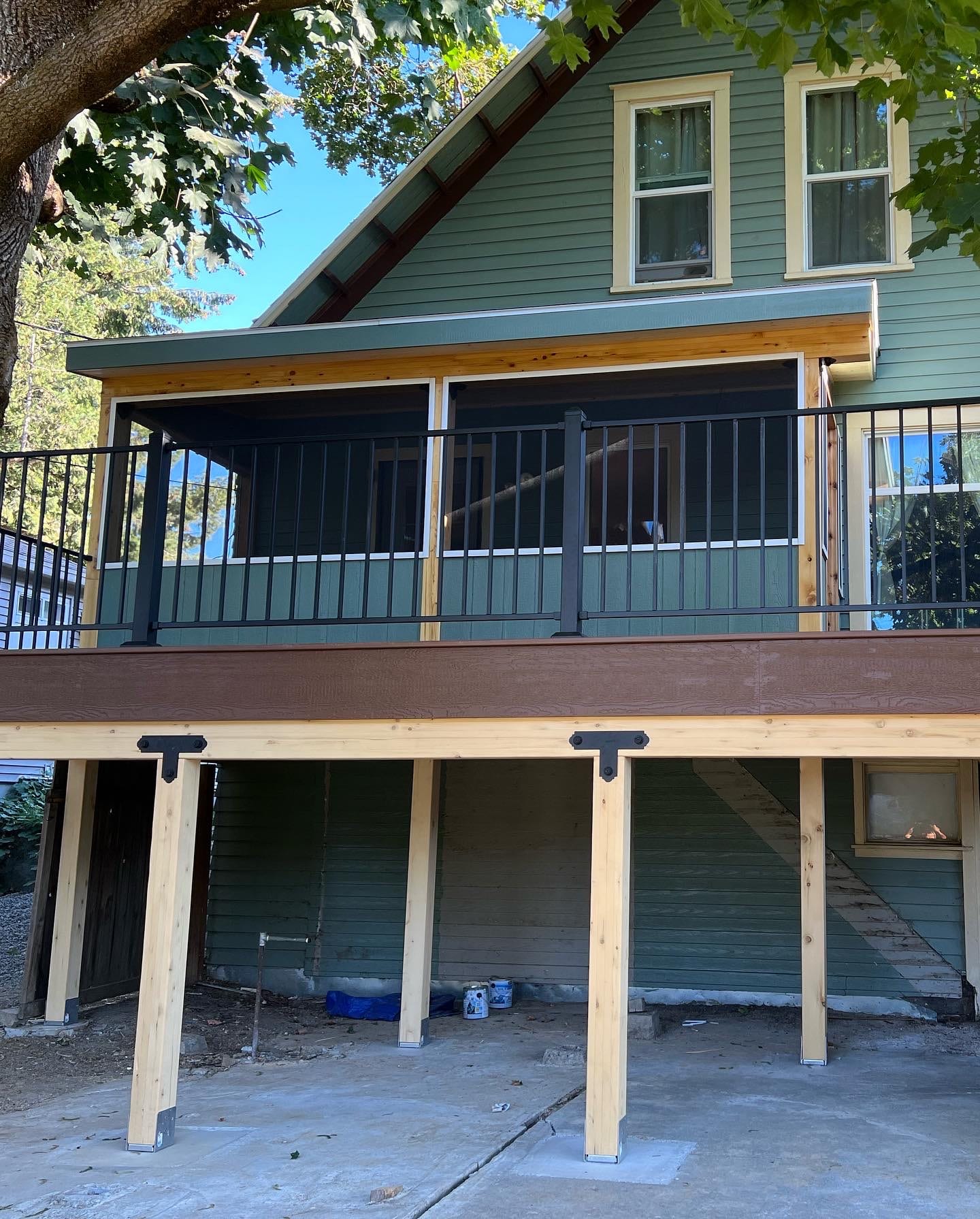 Green house with elevated screened porch above deck on stained timber posts, concrete pad beneath, tree overhead