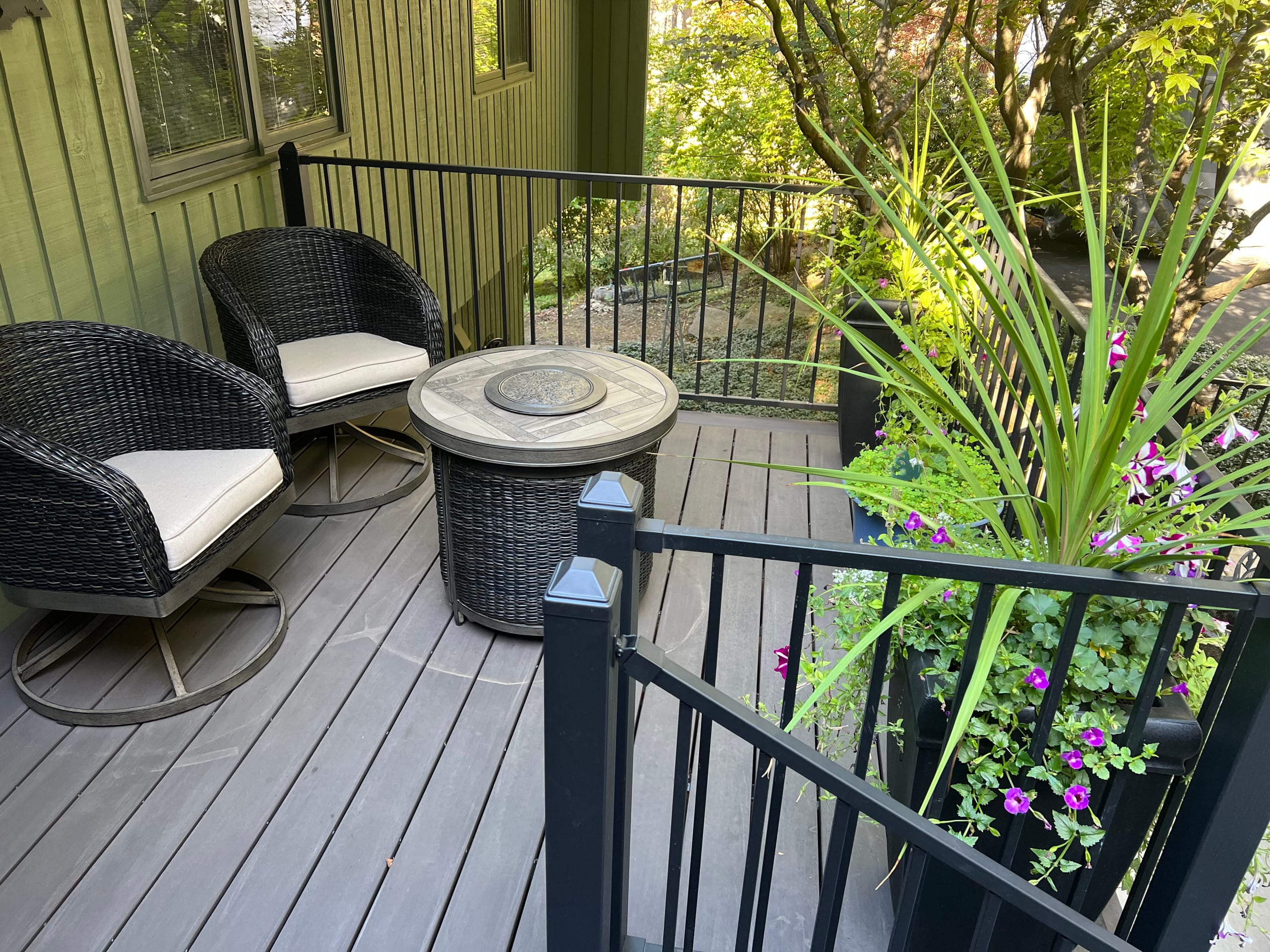 Grey-brown composite porch with black metal railing, wicker chairs, fire table and lush plants against green siding