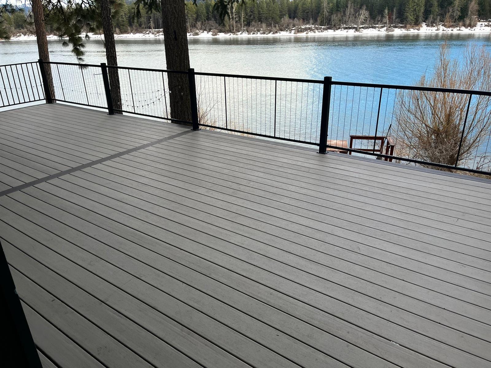 Grey composite deck with black cable railing right at a lake shore with snow patches