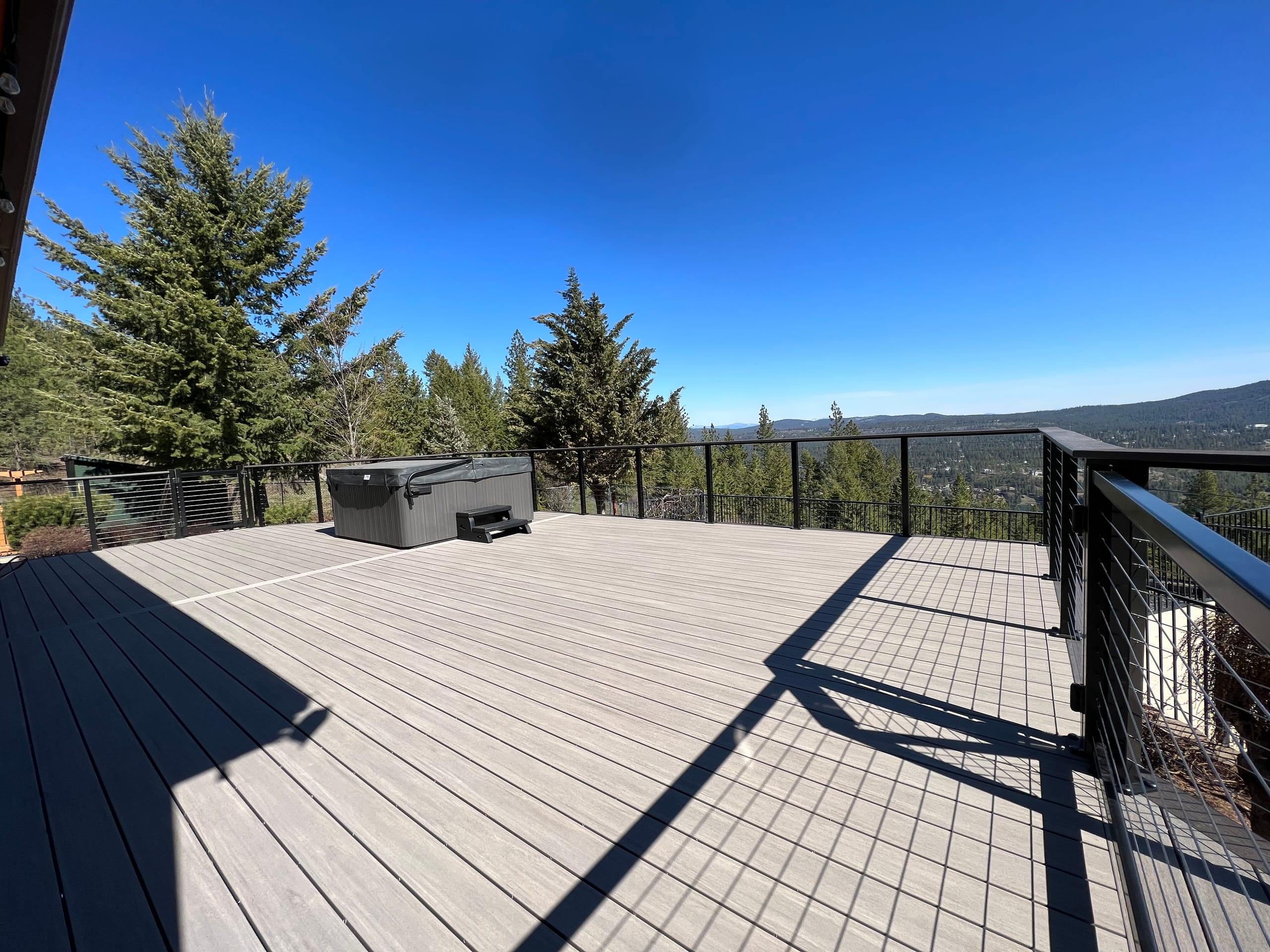 Grey composite deck with hot tub, cable railing and panoramic lake and hill view under deep blue sky