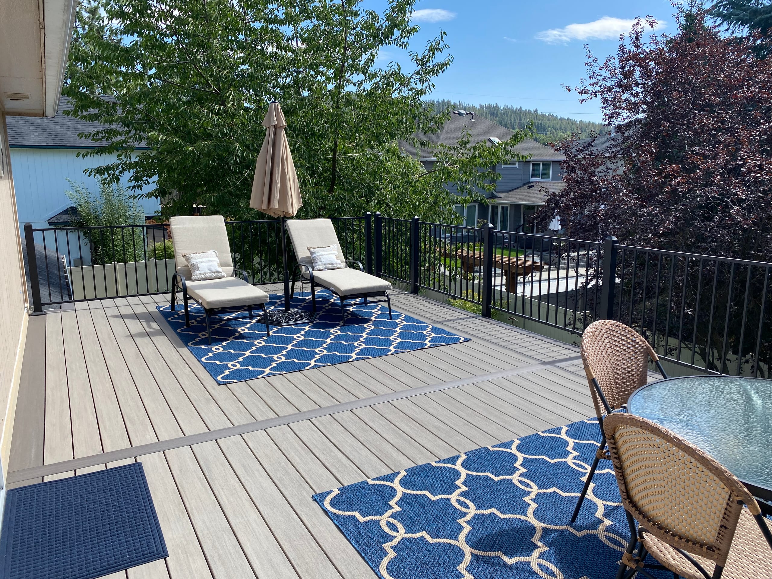 Grey composite deck with lounge chairs, umbrella and blue rug beside black railing and treetop view