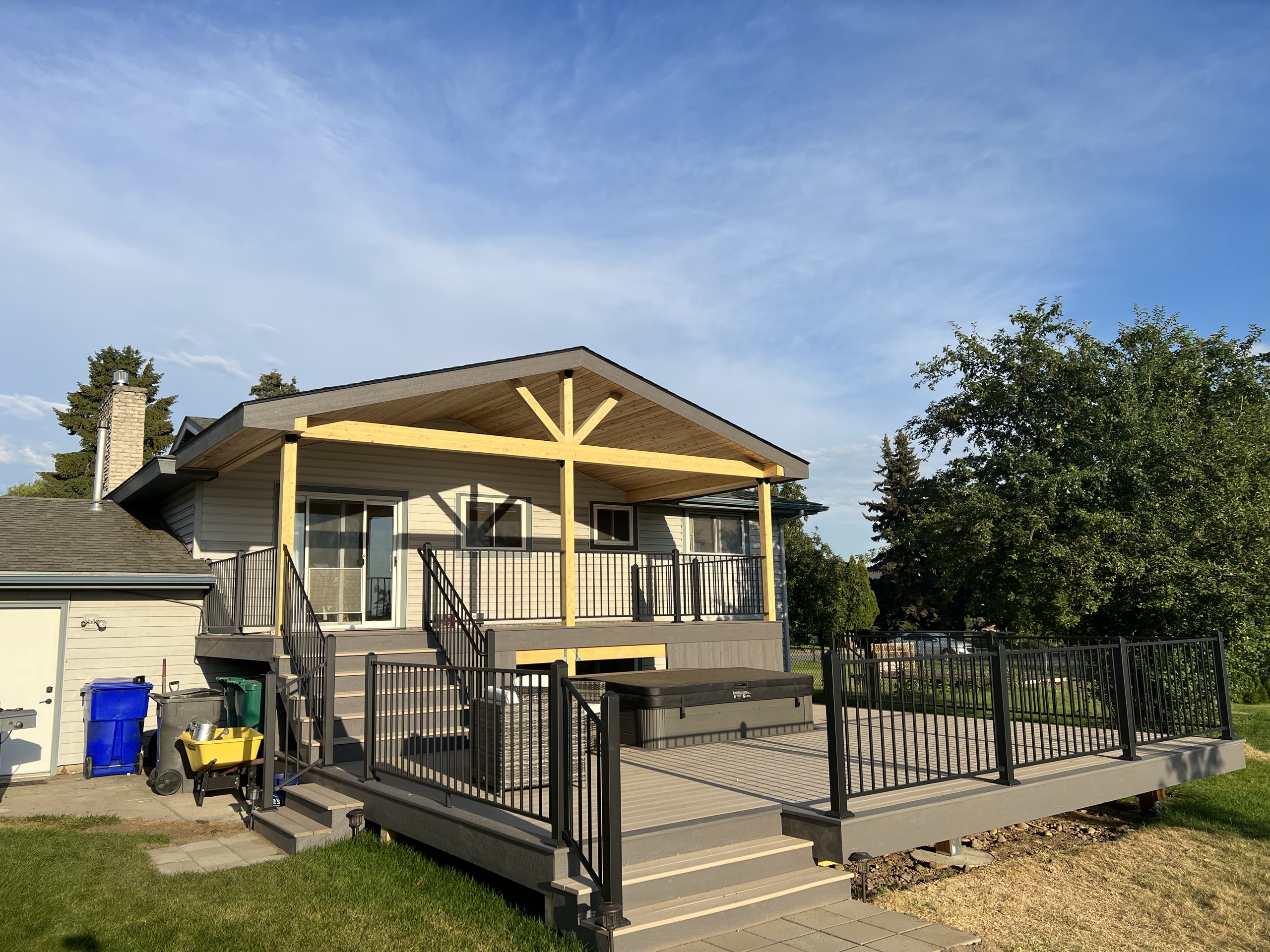 Grey composite deck with step lights, black railing, hot tub and gabled yellow-cedar patio cover on a tan house