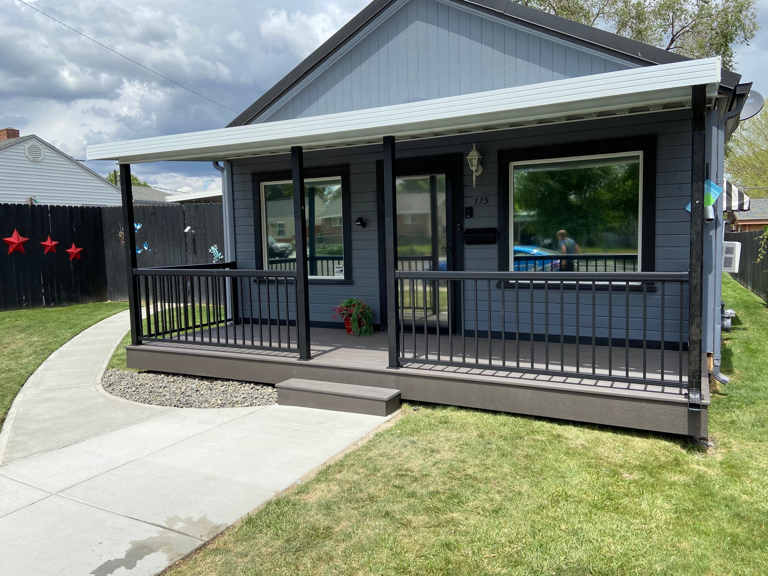 Grey cottage with covered front porch, dark composite deck, black metal picket railing and white aluminum roof