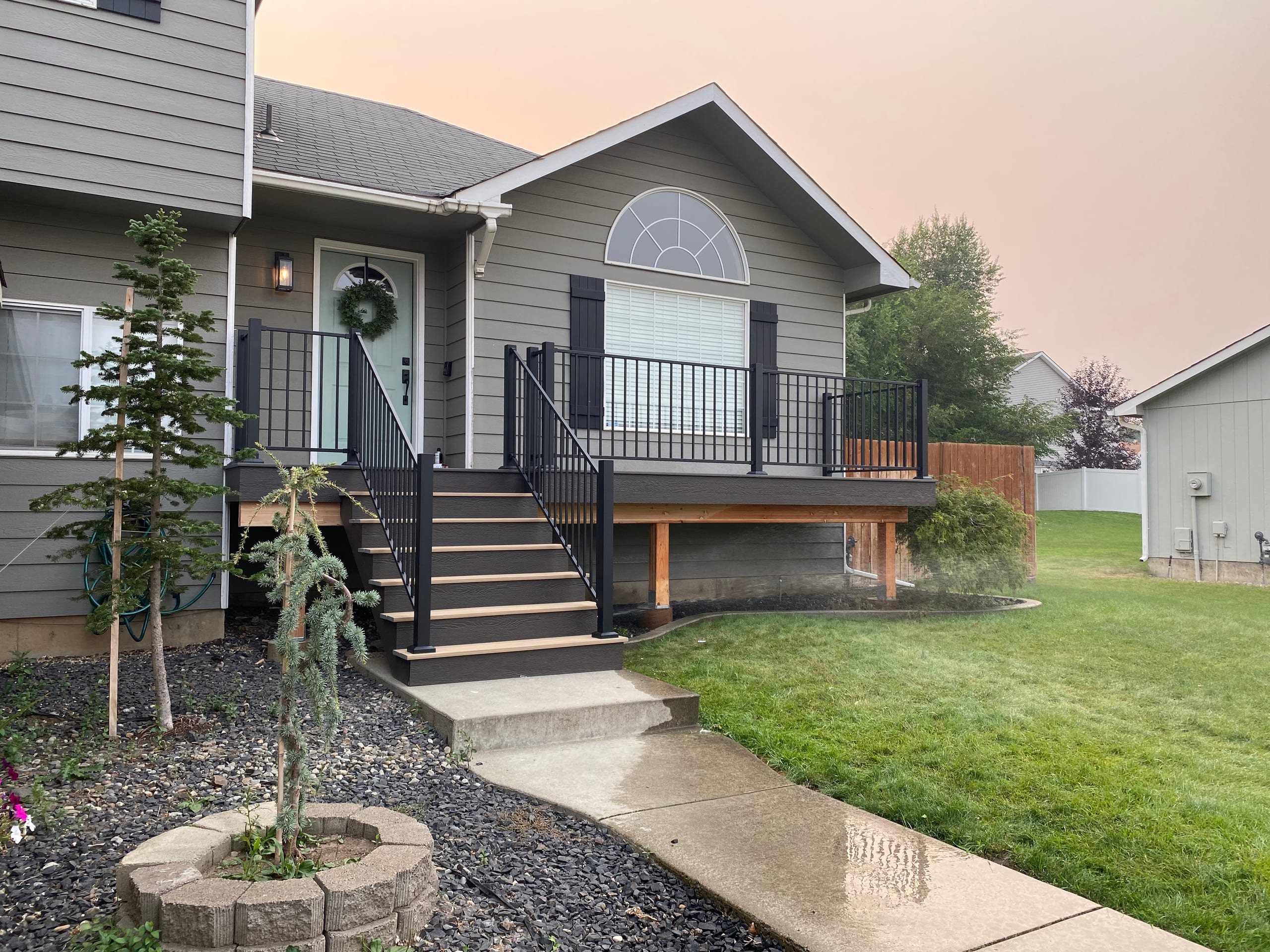 Grey house at dusk with elevated entry porch, dark composite stairs, black railing and wood trim, lawn and hazy pink sky