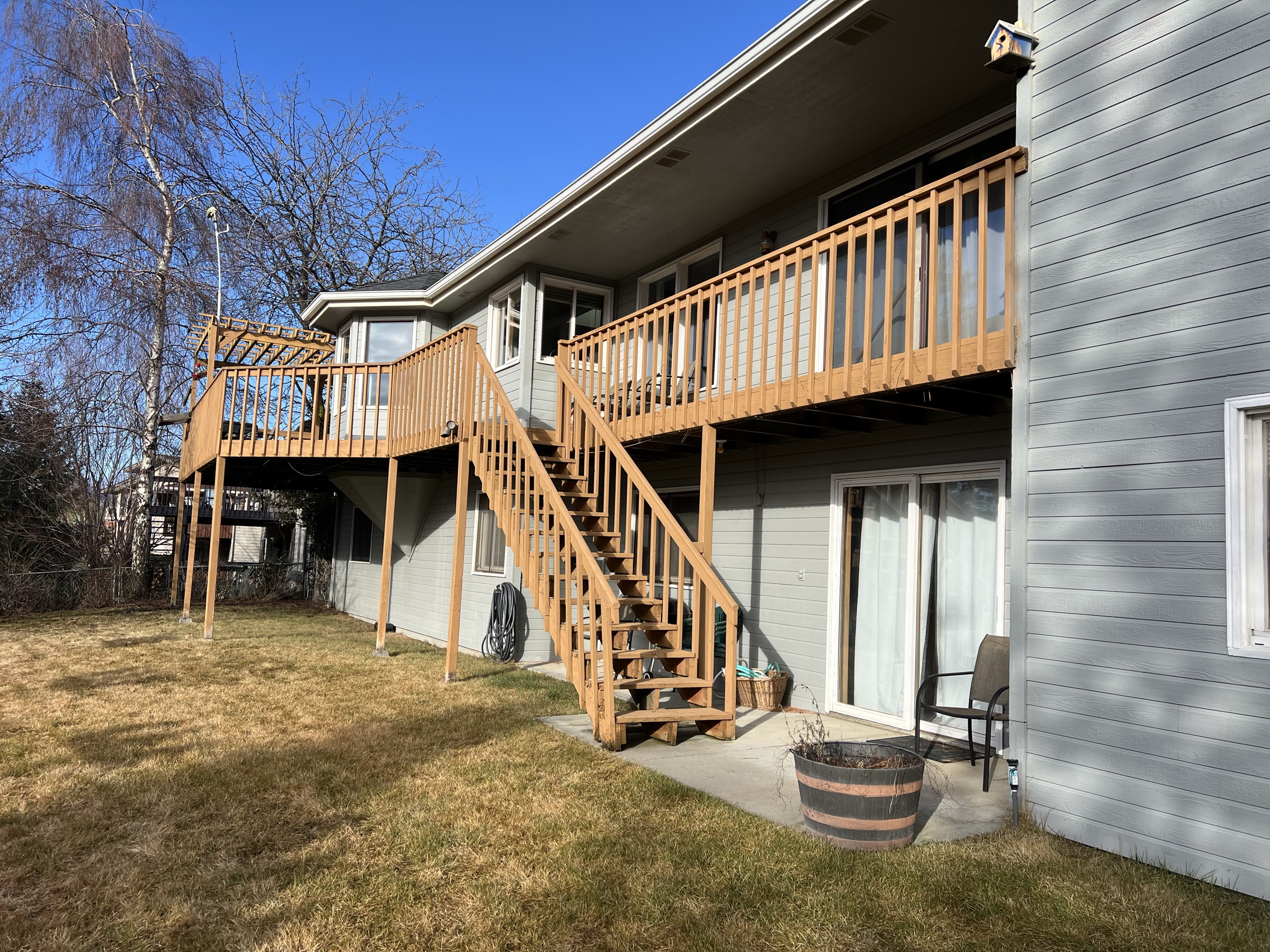 Grey house with elevated cedar-stained wood deck, wood railing and long wood stair, pergola corner, dry lawn