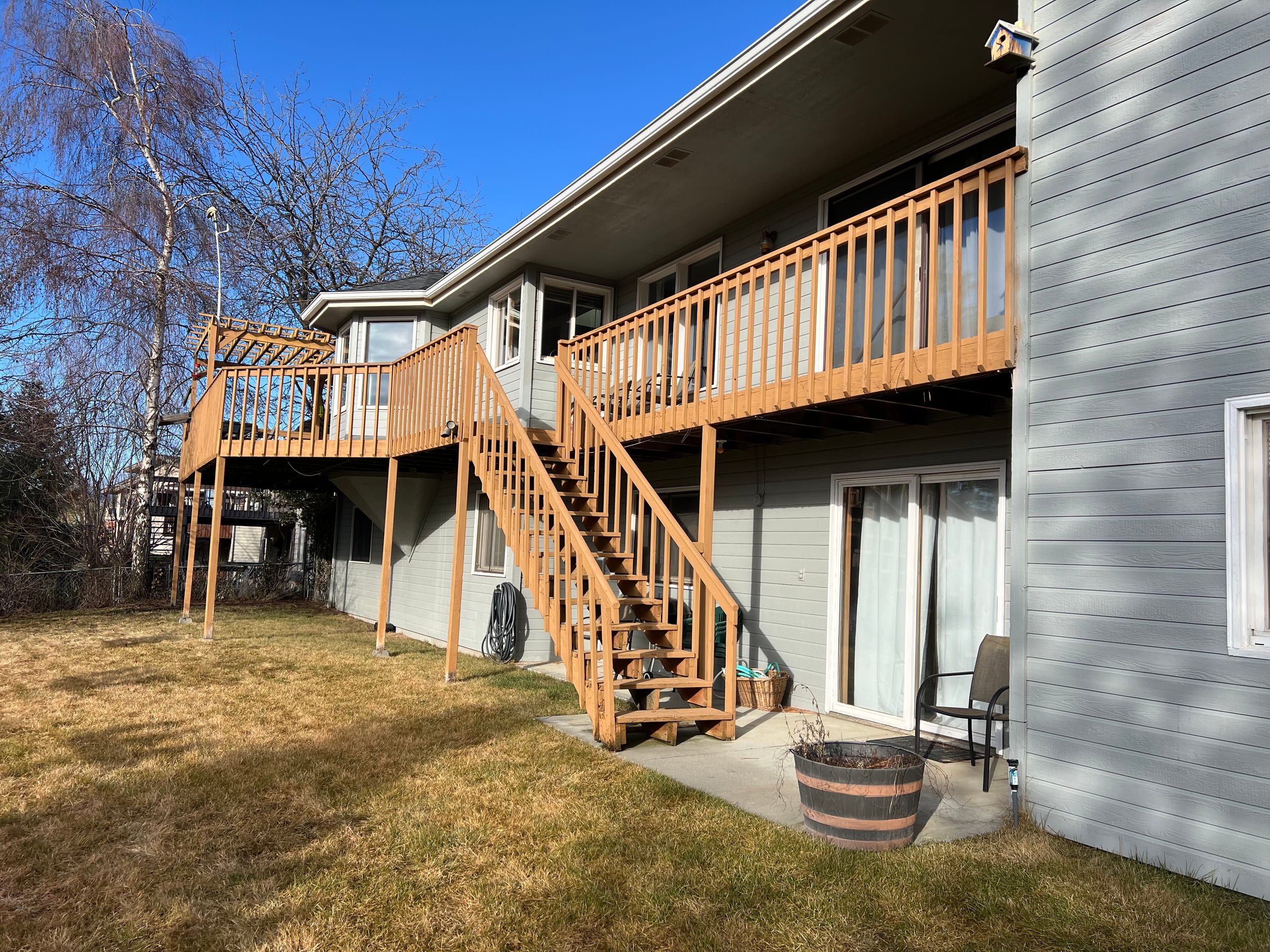 Grey house with elevated cedar-stained wood deck, wood railing and long wood stair, pergola corner, dry lawn