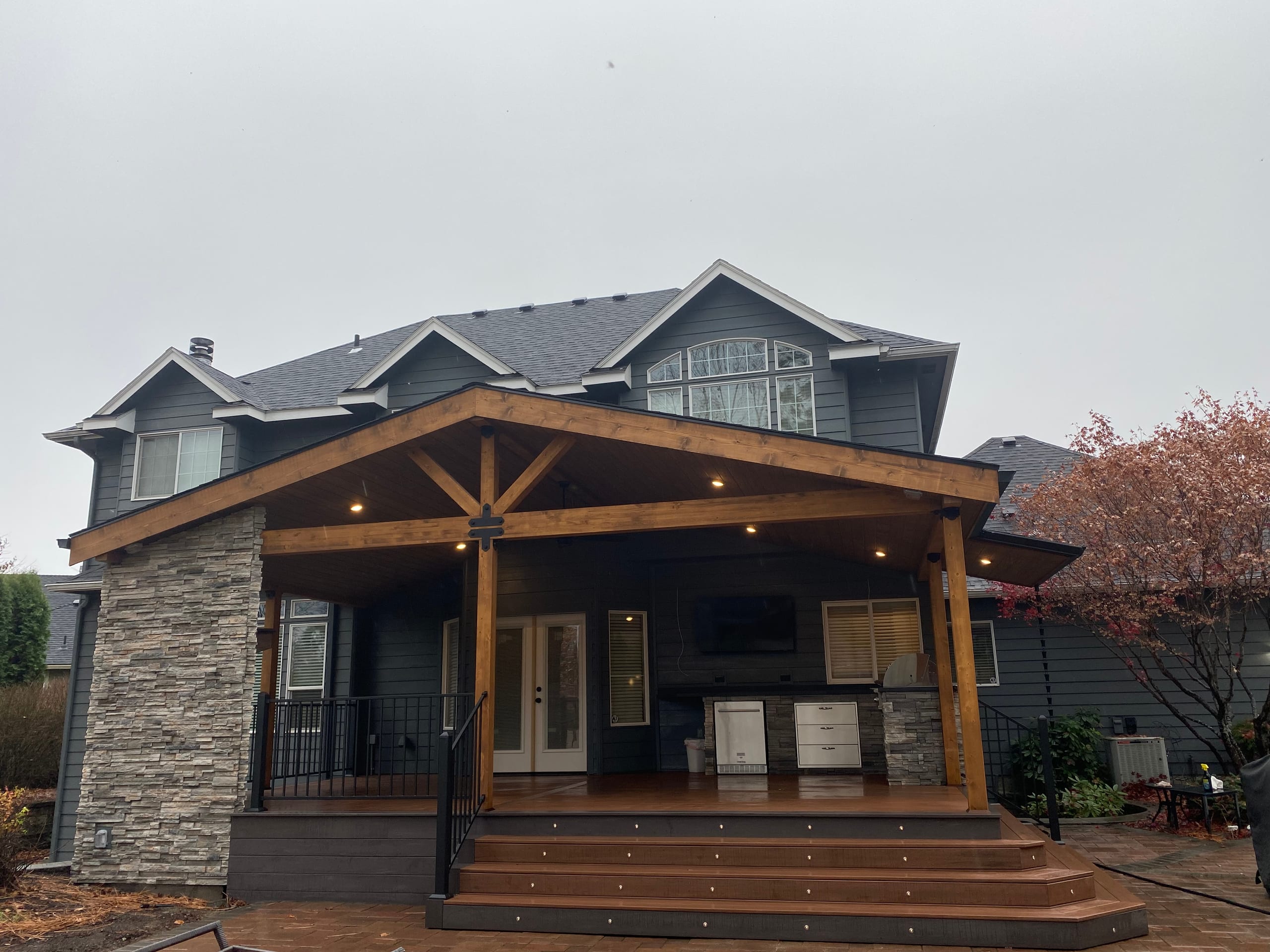 Head-on view of gable covered deck with stone pier, step lights, stained trusses on a gray house, overcast day