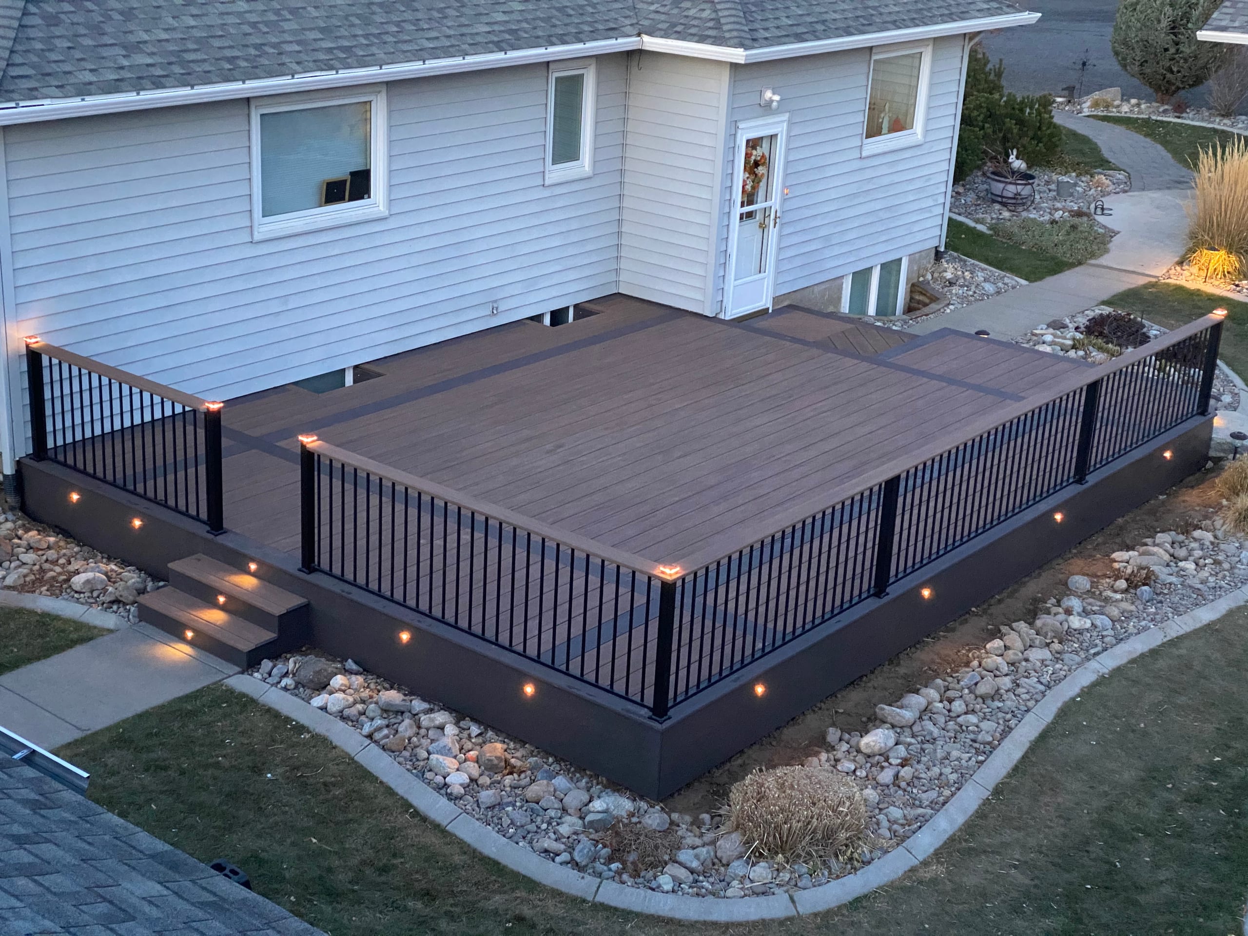 High-angle view of dark composite deck with black picket railing, lit steps and post caps beside pale house