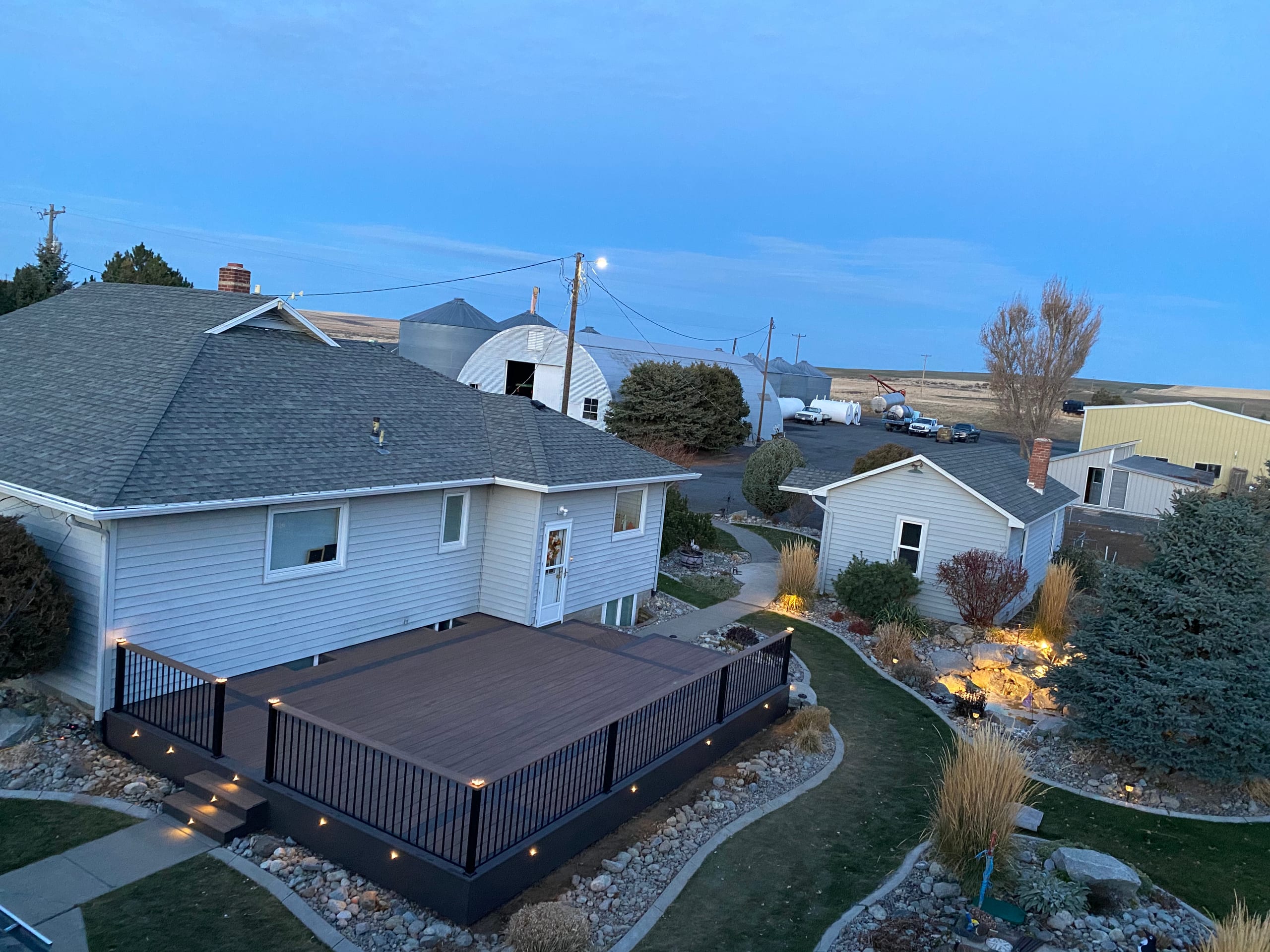 High-angle dusk view of dark composite deck with black picket railing and accent lighting beside pale house and rock garden