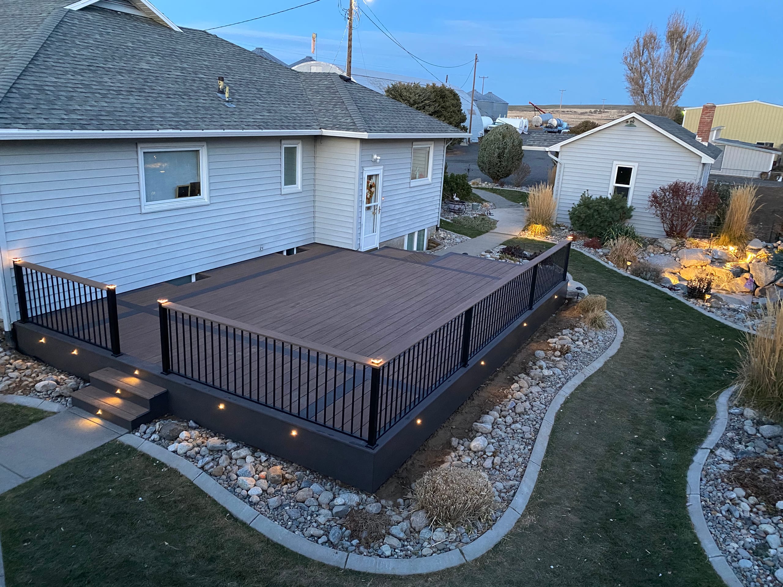 High-angle dusk view of dark composite deck with black picket railing, lit steps and rock-lined lawn