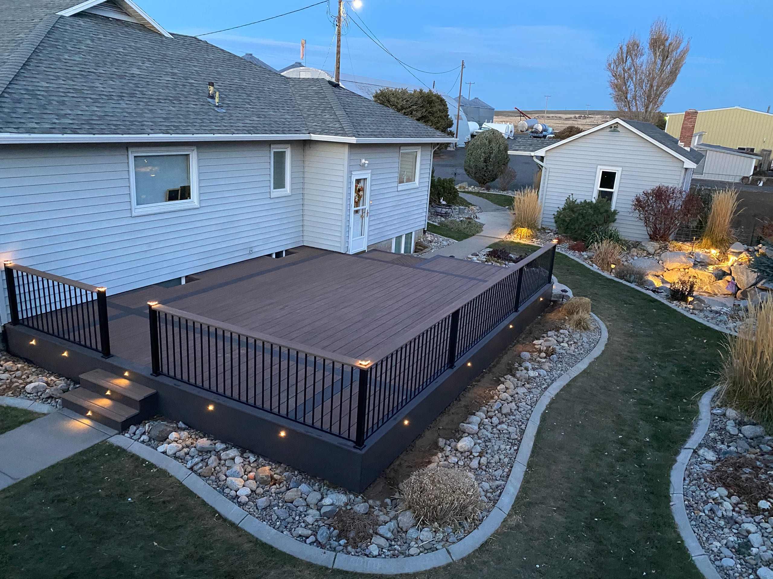 High-angle dusk view of dark composite deck with black picket railing, lit steps and rock garden beside pale house
