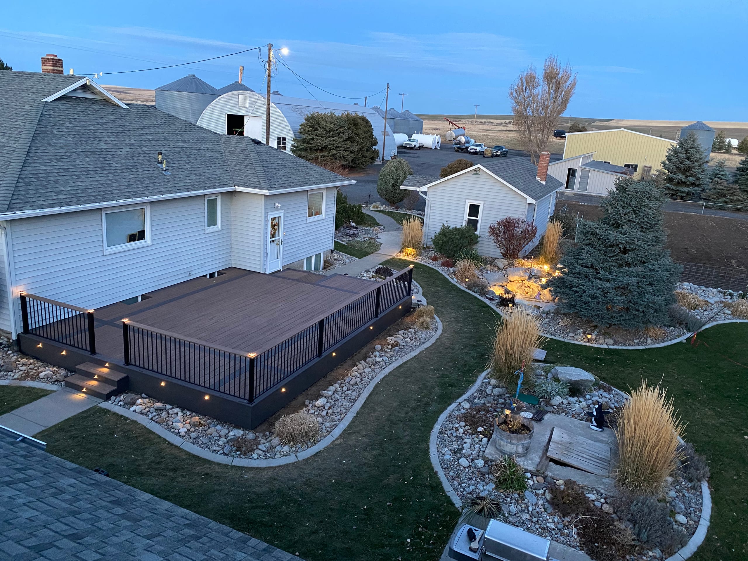 High-angle dusk view of low dark deck with railing in landscaped yard, outbuildings behind