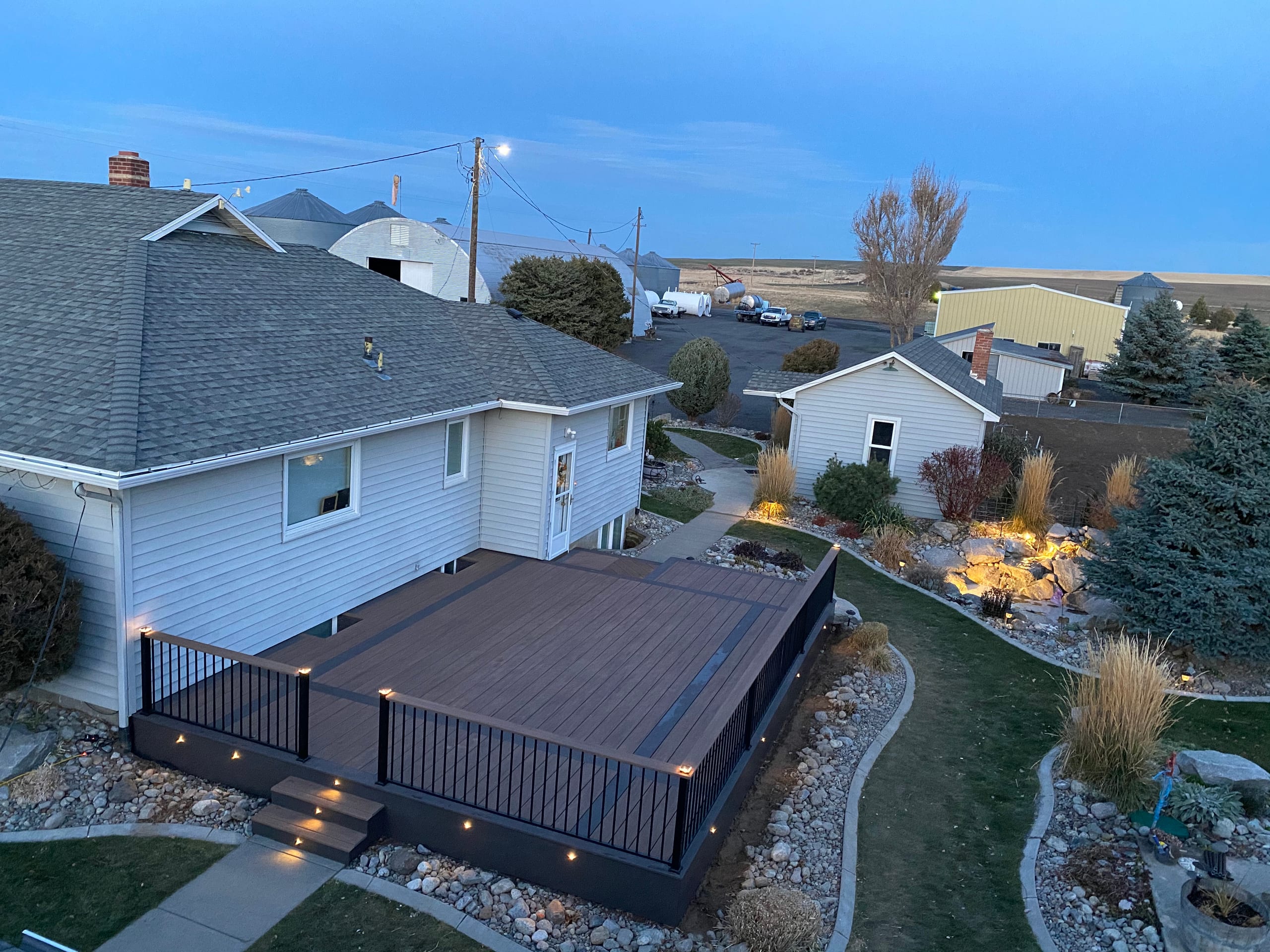 High-angle view of finished dark composite deck with black railing and lights in a landscaped backyard at dusk
